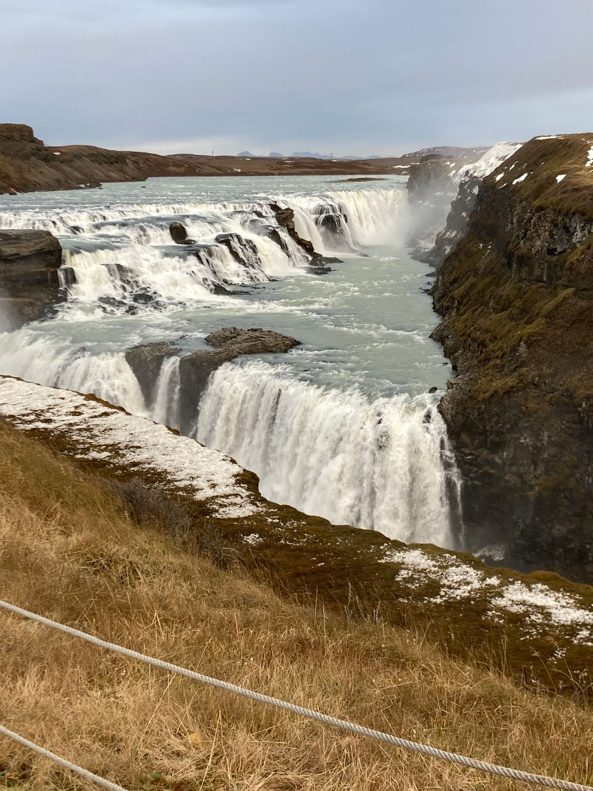 Waterfall cascading down multiple tiers into a river. Snow on the banks, overcast sky.