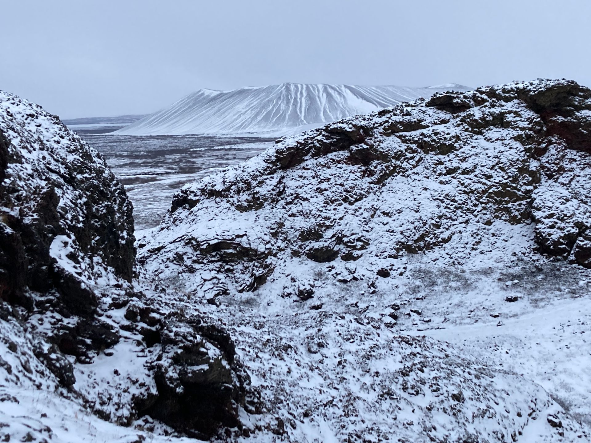 Snowy volcanic landscape with a distant, ridged mountain under a cloudy sky.