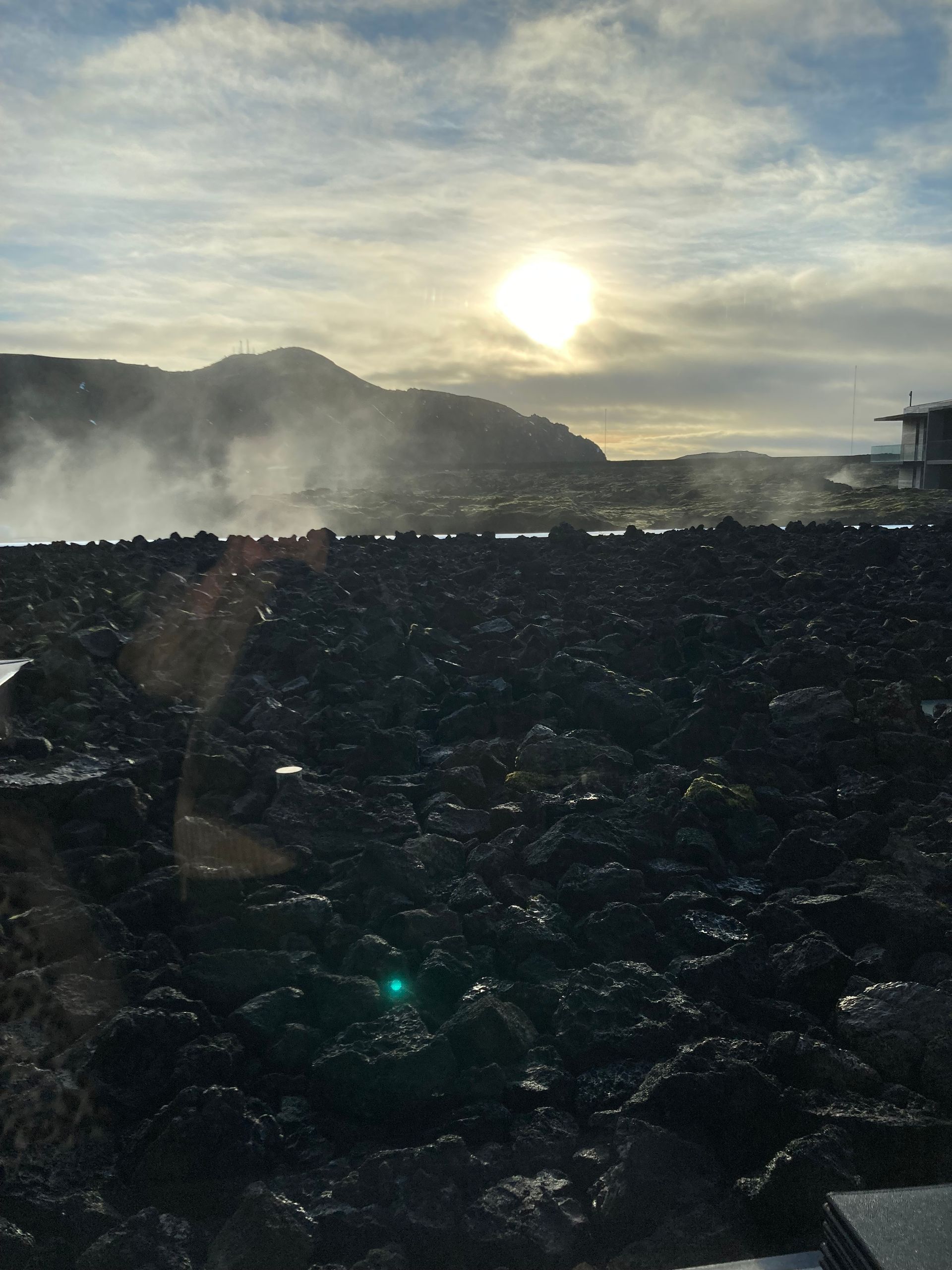 Sun shines over black lava rocks, steam rising near a mountain in Iceland.