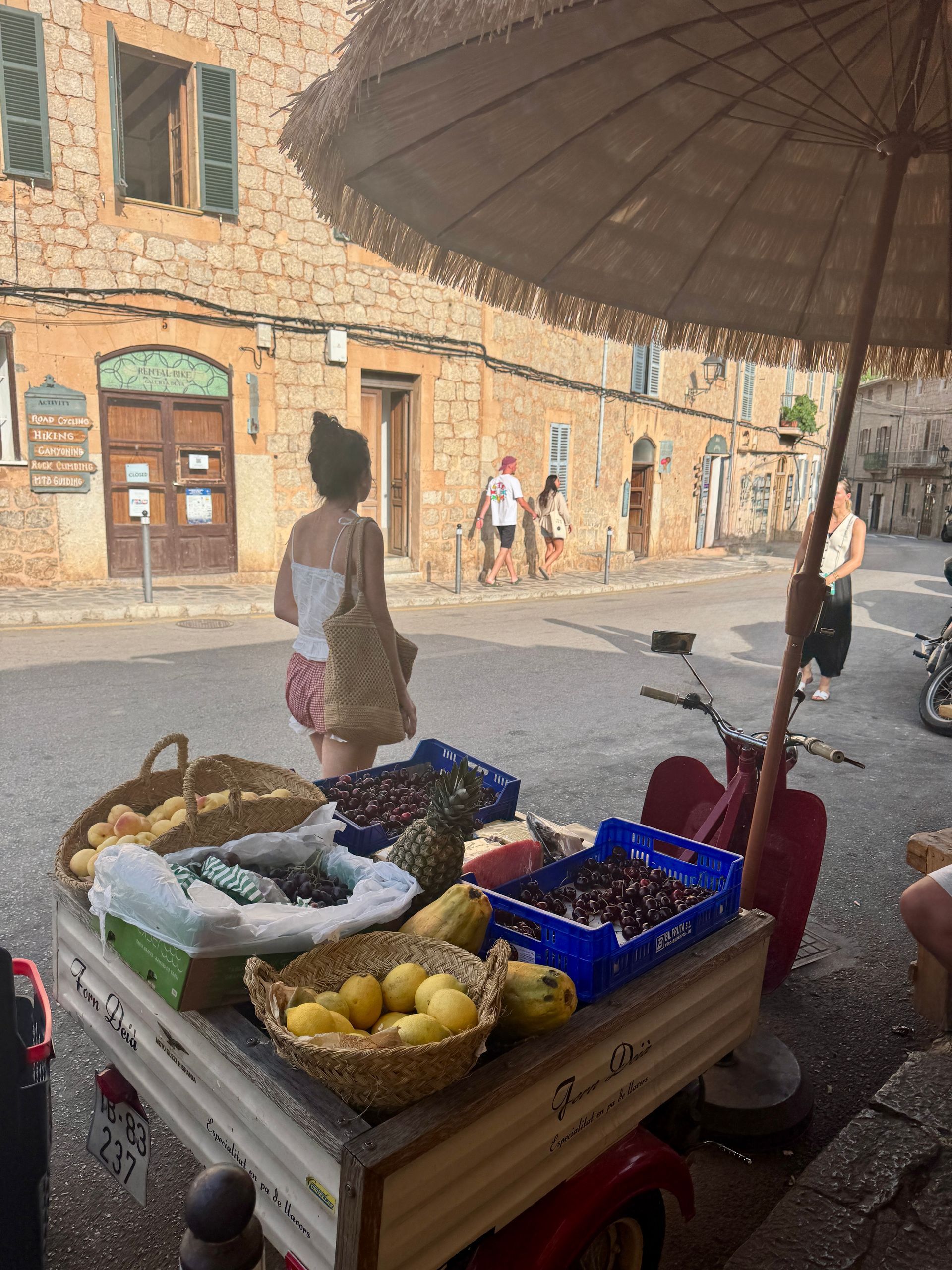 Fruit stand on a street in front of a stone building. A person with a basket is near the stand.