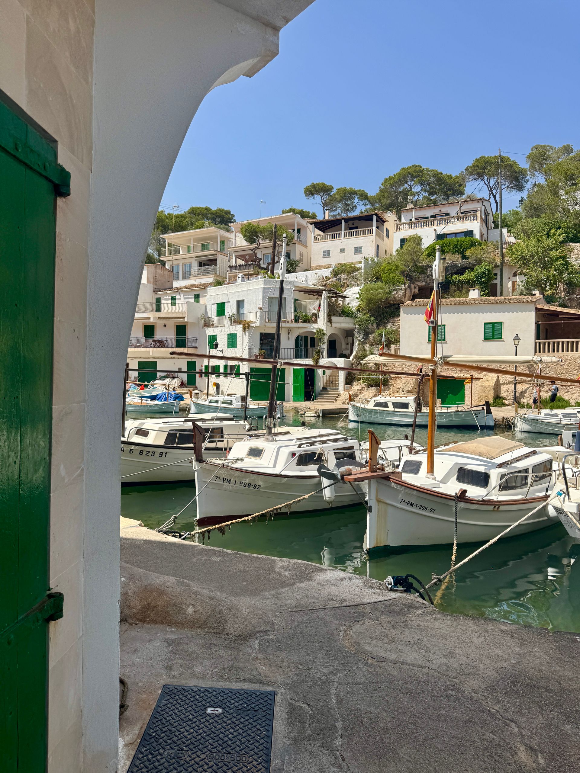 Harbor view through a white archway, boats docked, white buildings on the hillside, blue sky.