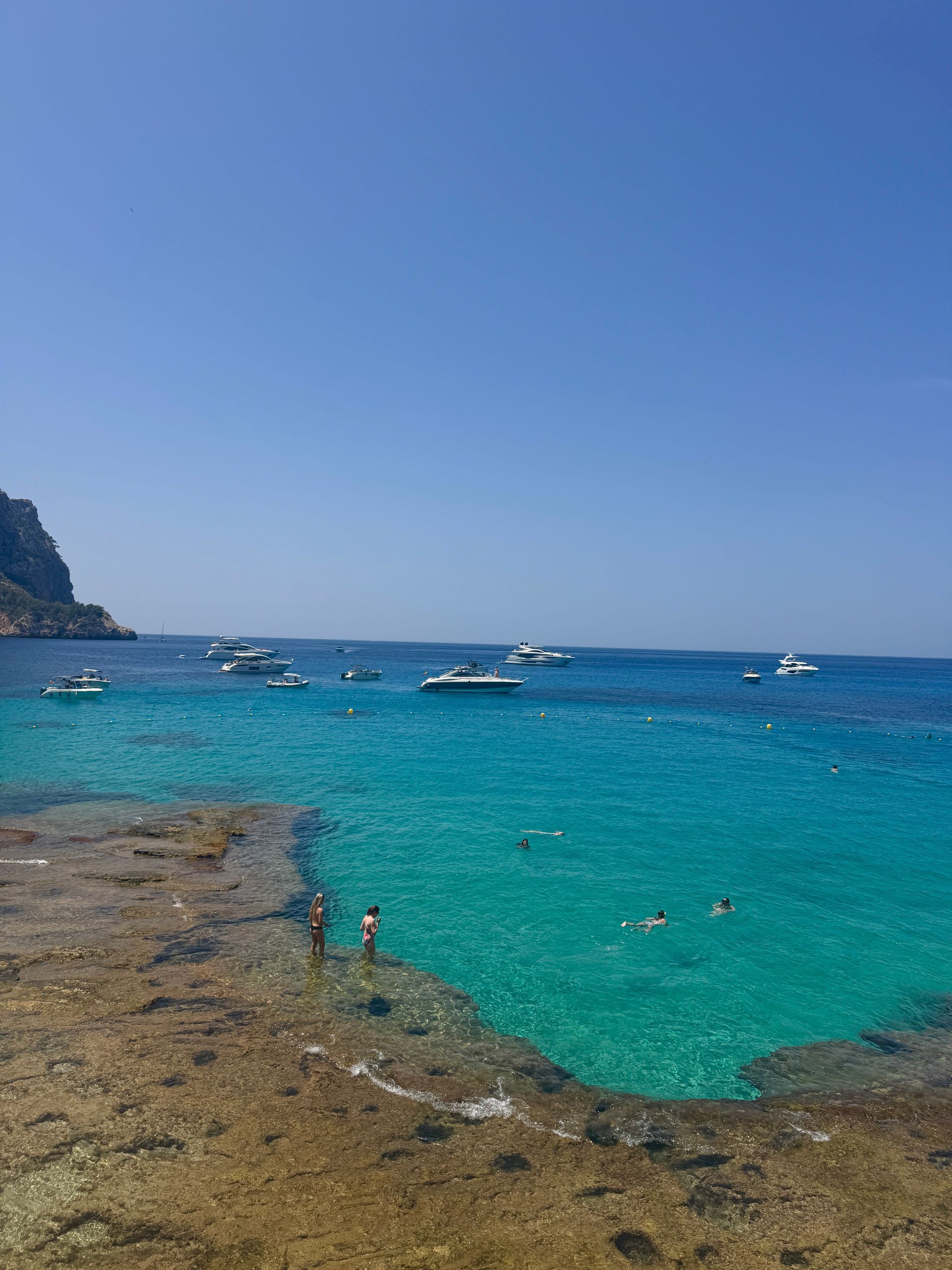People swimming in turquoise water near boats, cliffs under blue sky in Majorca, Spain.