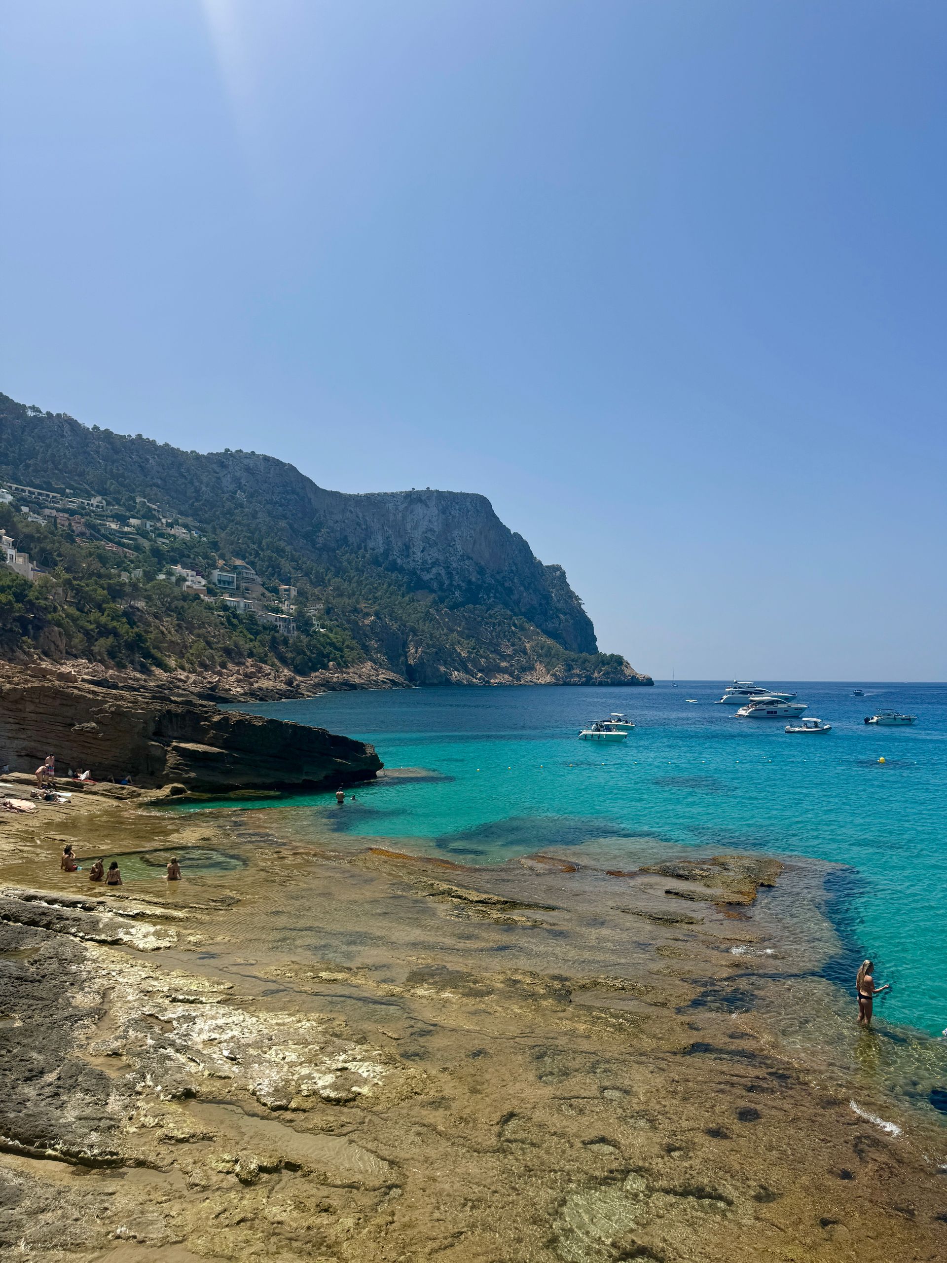 Coastal view: turquoise water, rocky shore, boats, cliffside, clear blue sky.