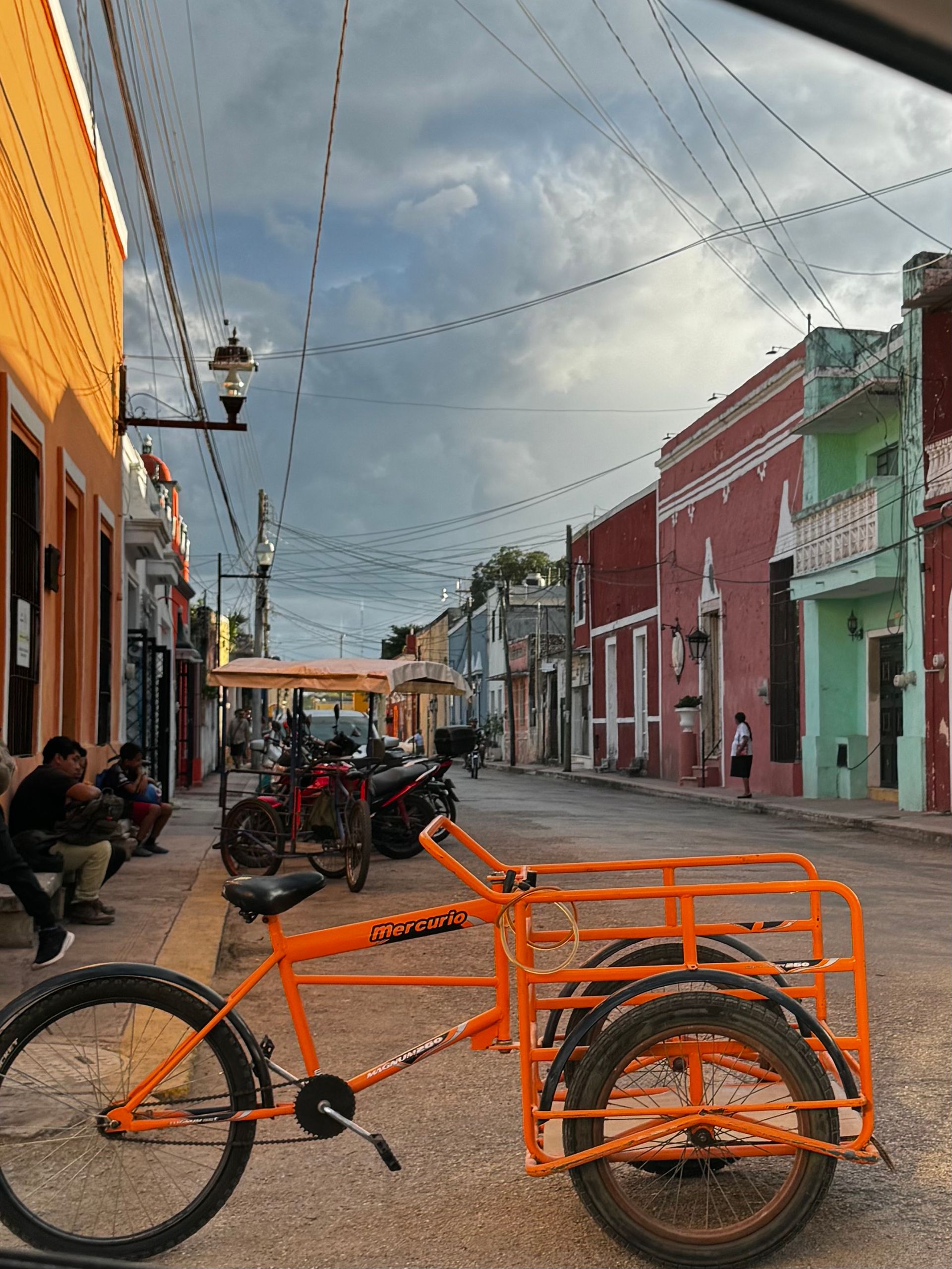 A cobblestone street lined with colorful buildings. People are sitting on a bench in front in Valladolid, Mexico.
