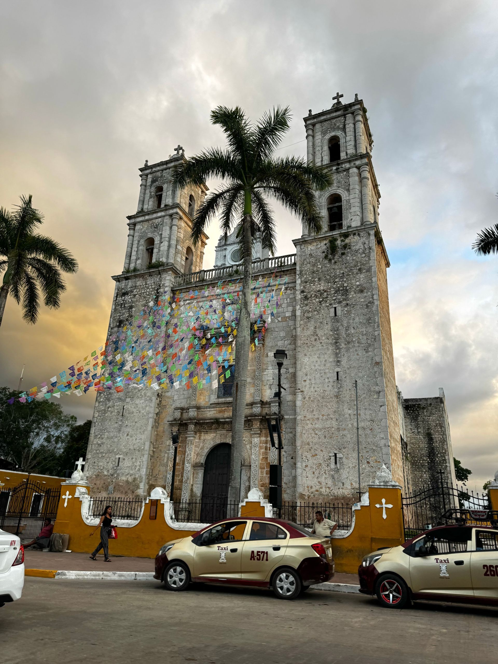 Old stone church with bell towers, palm tree, colorful banners, and parked cars under cloudy sky.