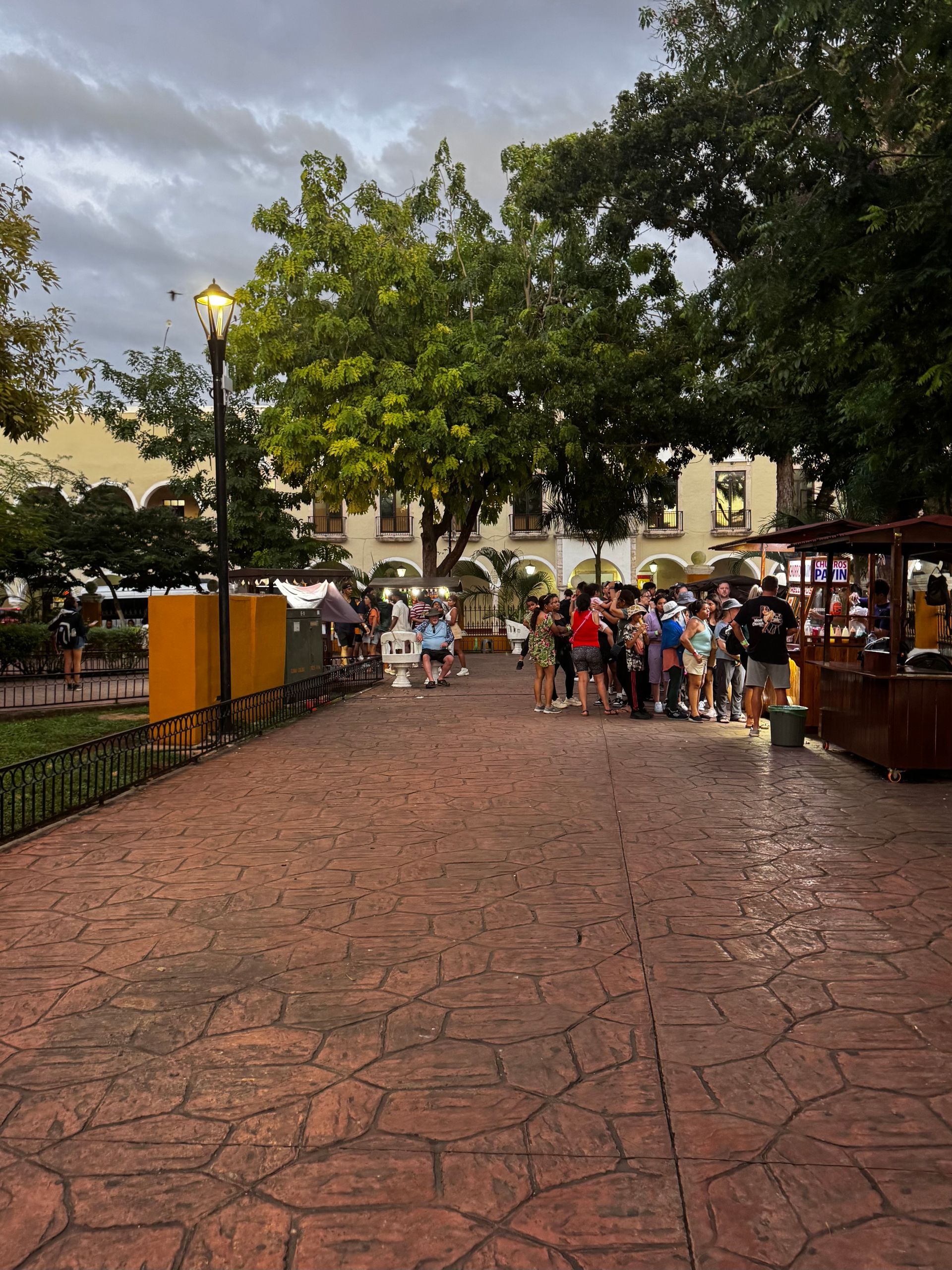 People line up at food stall in a plaza with trees and buildings in the background in Valladolid, Mexico.