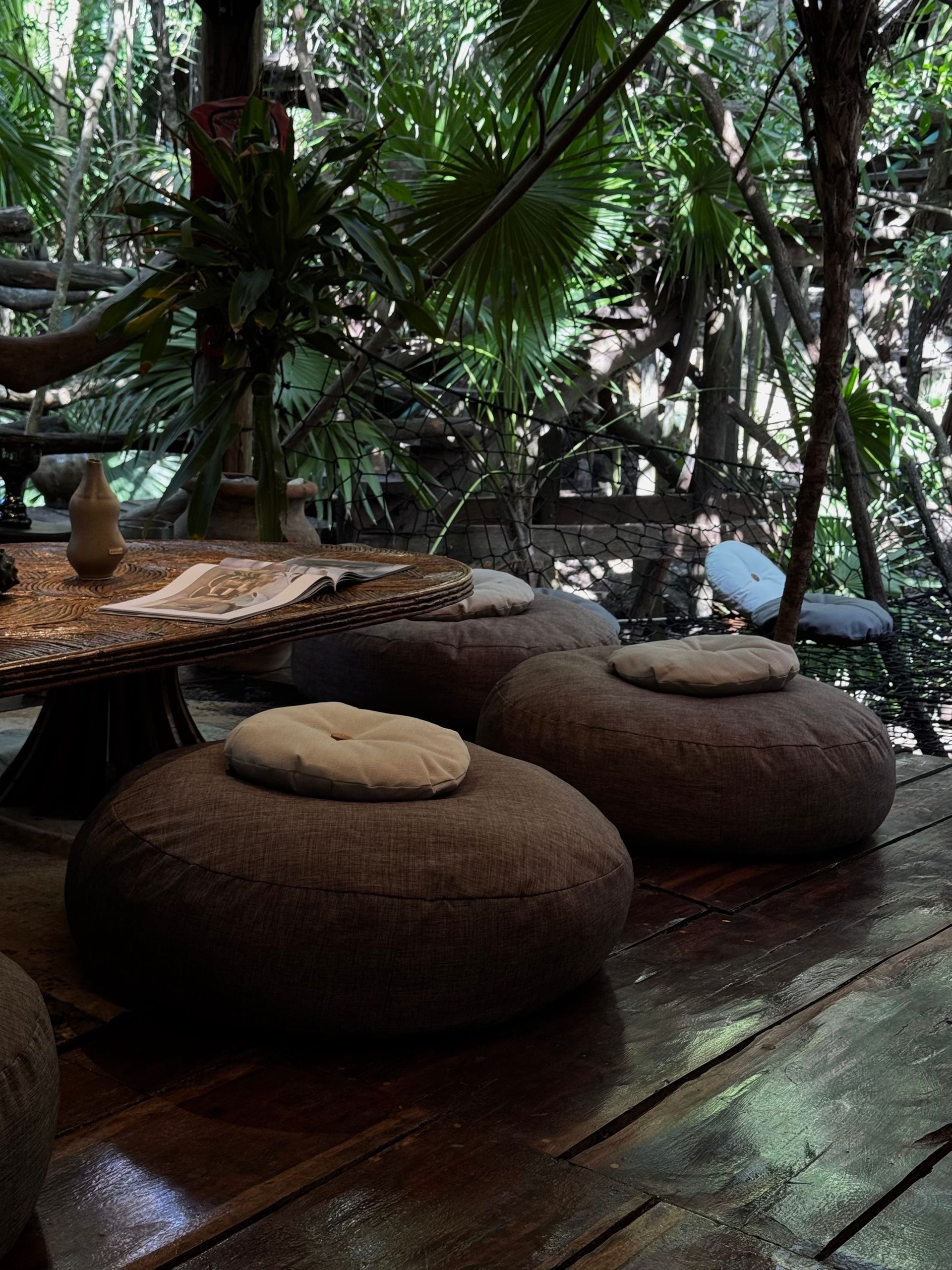 Wooden deck with round floor cushions and a table, surrounded by lush tropical foliage.