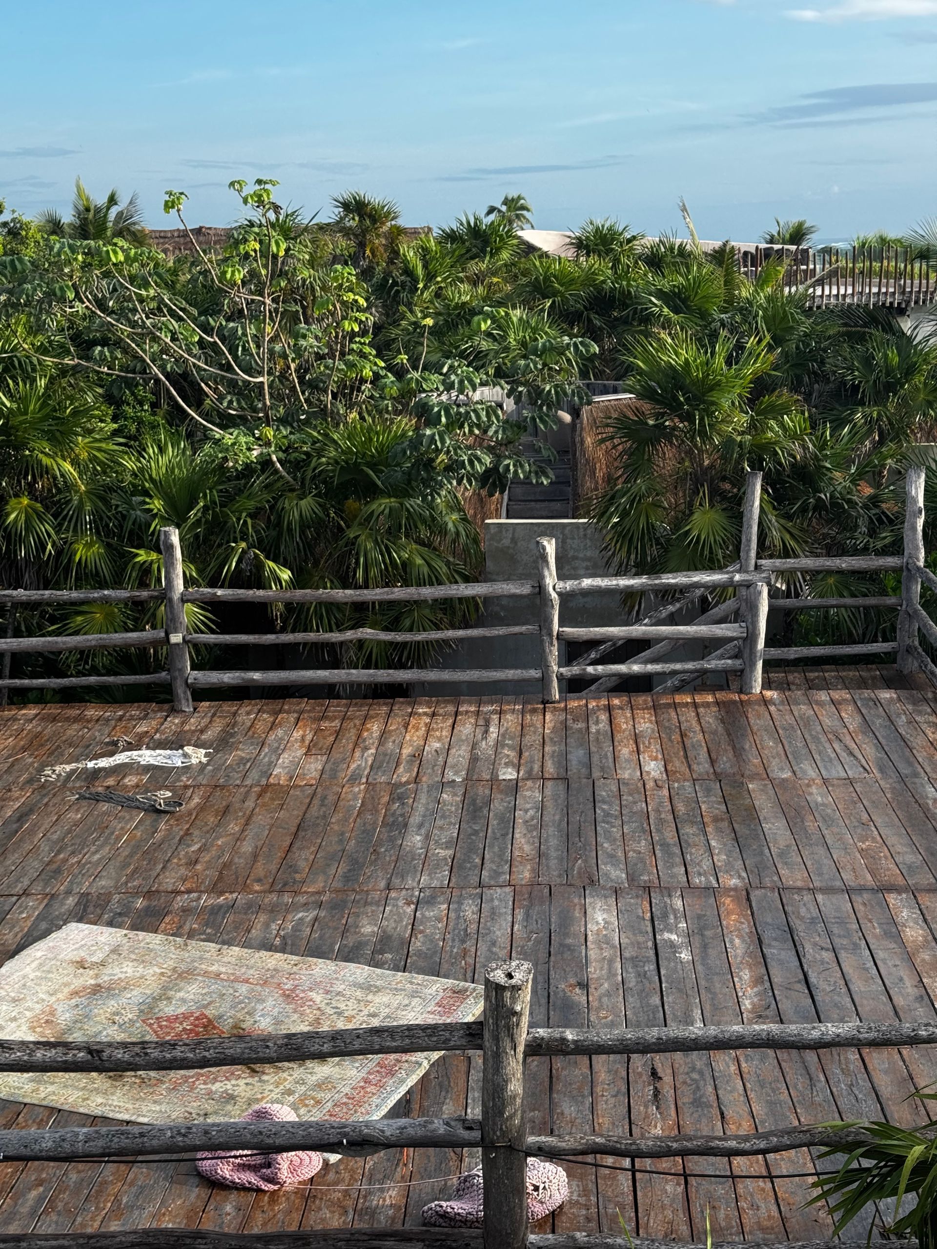 Wooden deck with a rustic fence overlooking lush green foliage under a blue sky in Tulum, Mexico.