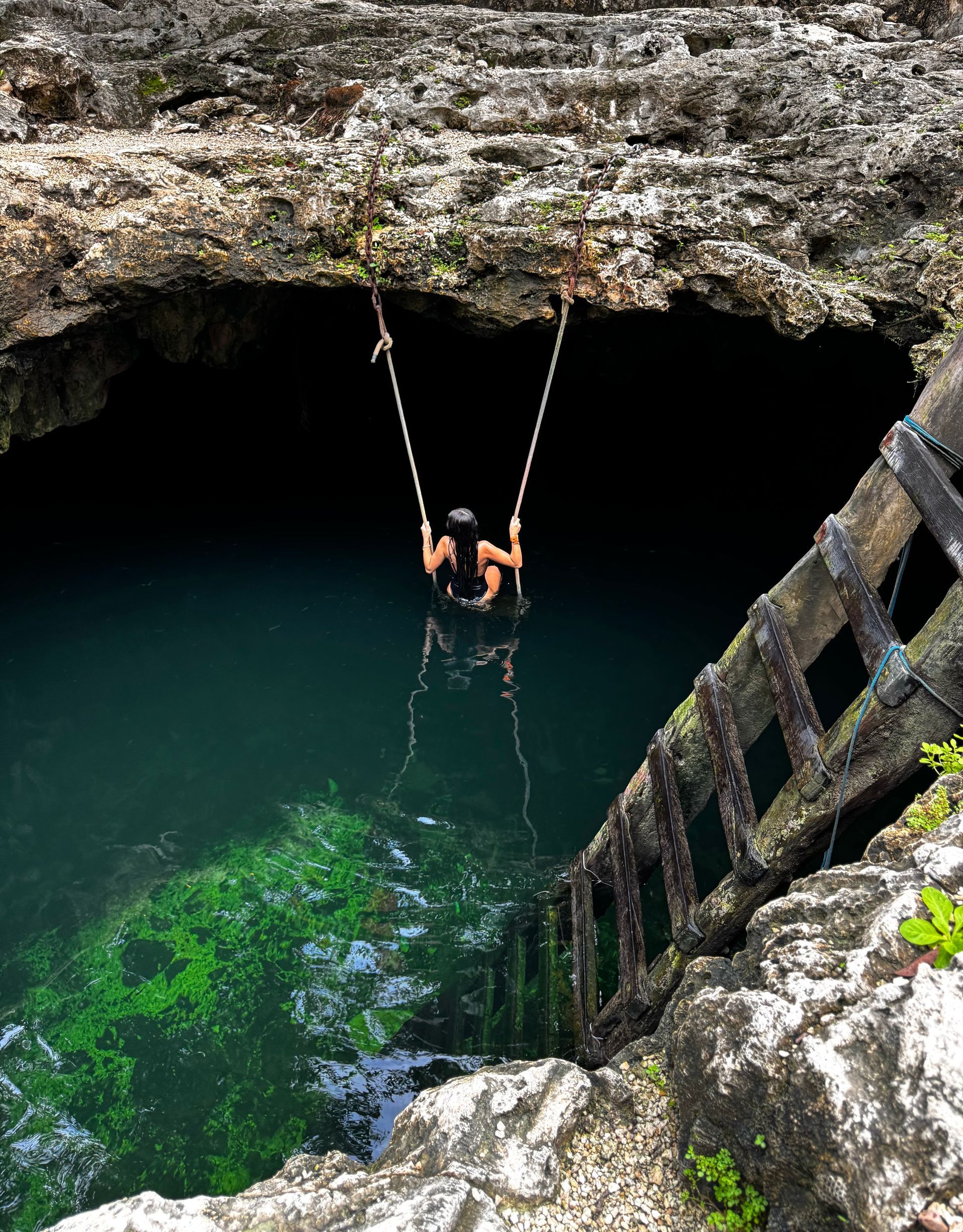 Person on a swing suspended over dark water inside a cavern. A weathered wooden ladder leads into the water in Tulum, Mexico.