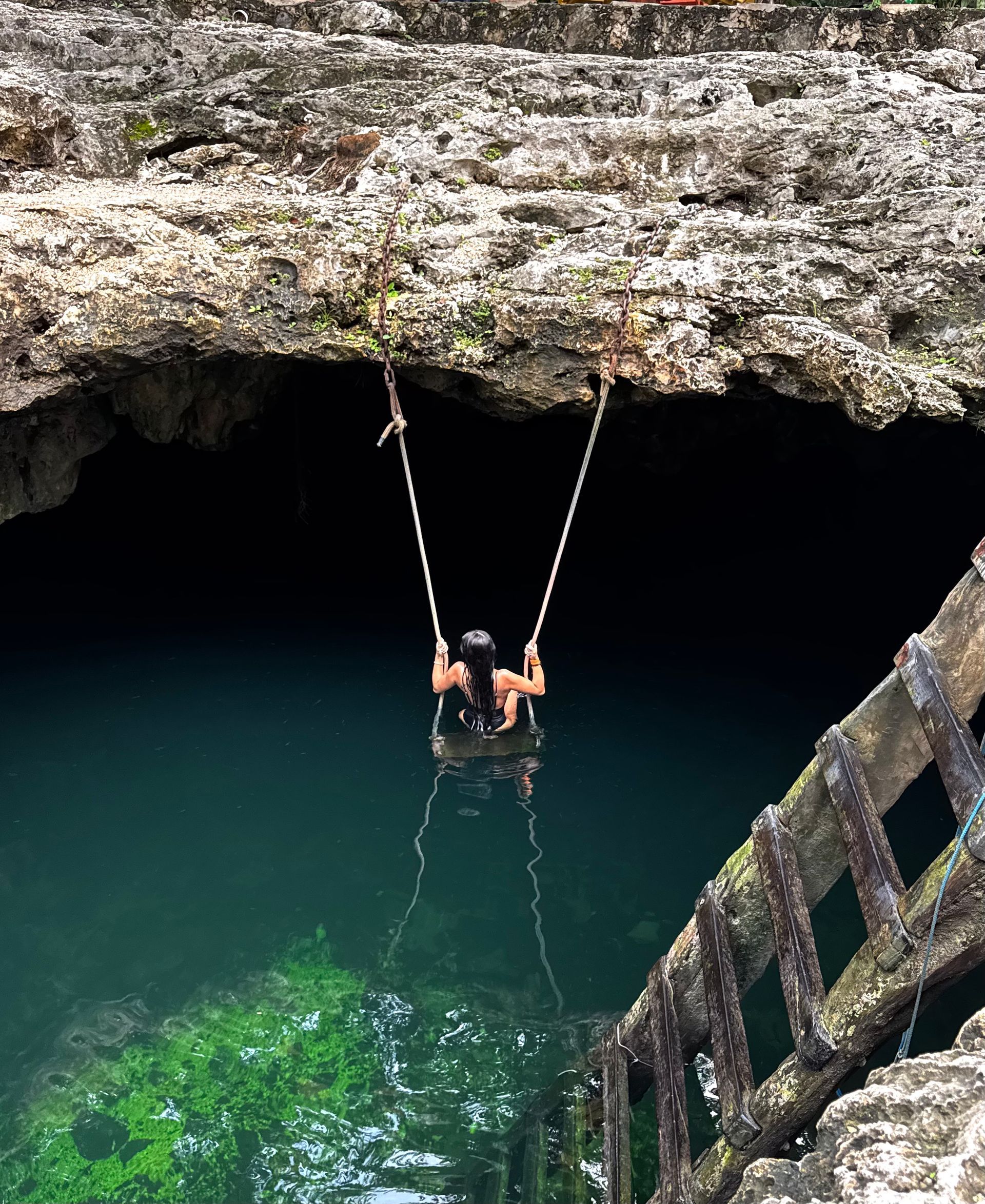 Person swings over dark blue water from a rope swing in a cavern.