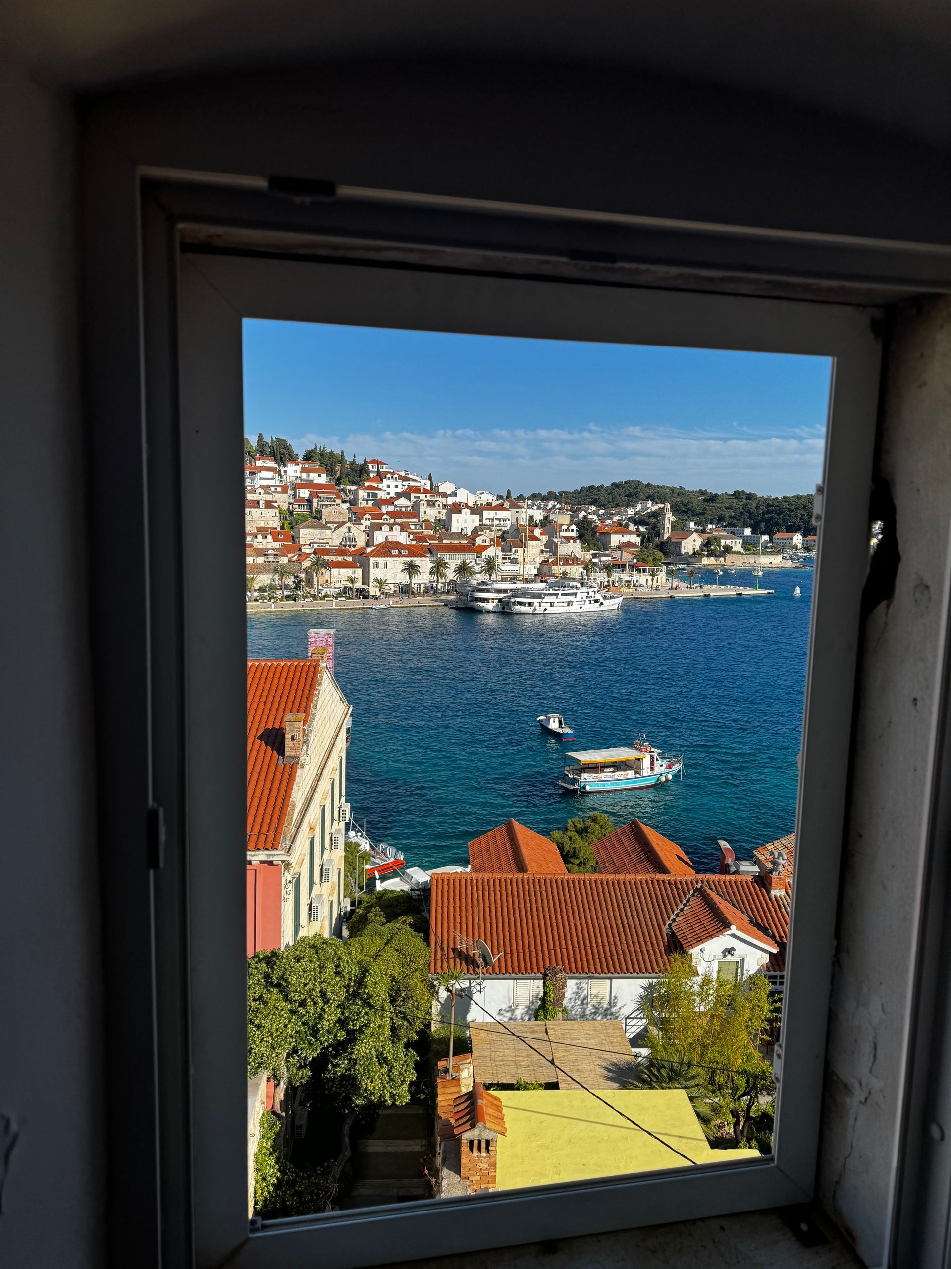 View of a coastal town through a window. Blue sea, red-roofed buildings, boats, and clear sky.