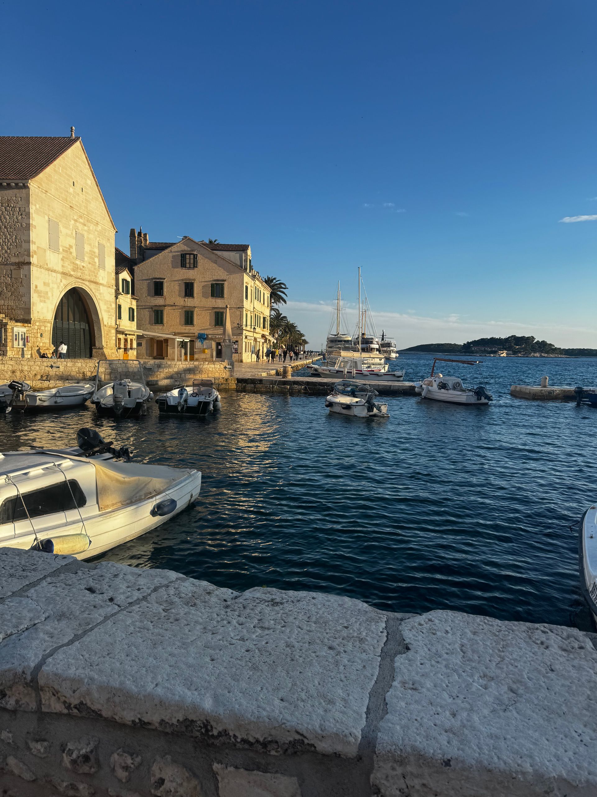 Boats in a harbor next to buildings under a blue sky. Sunlight reflects on the water.
