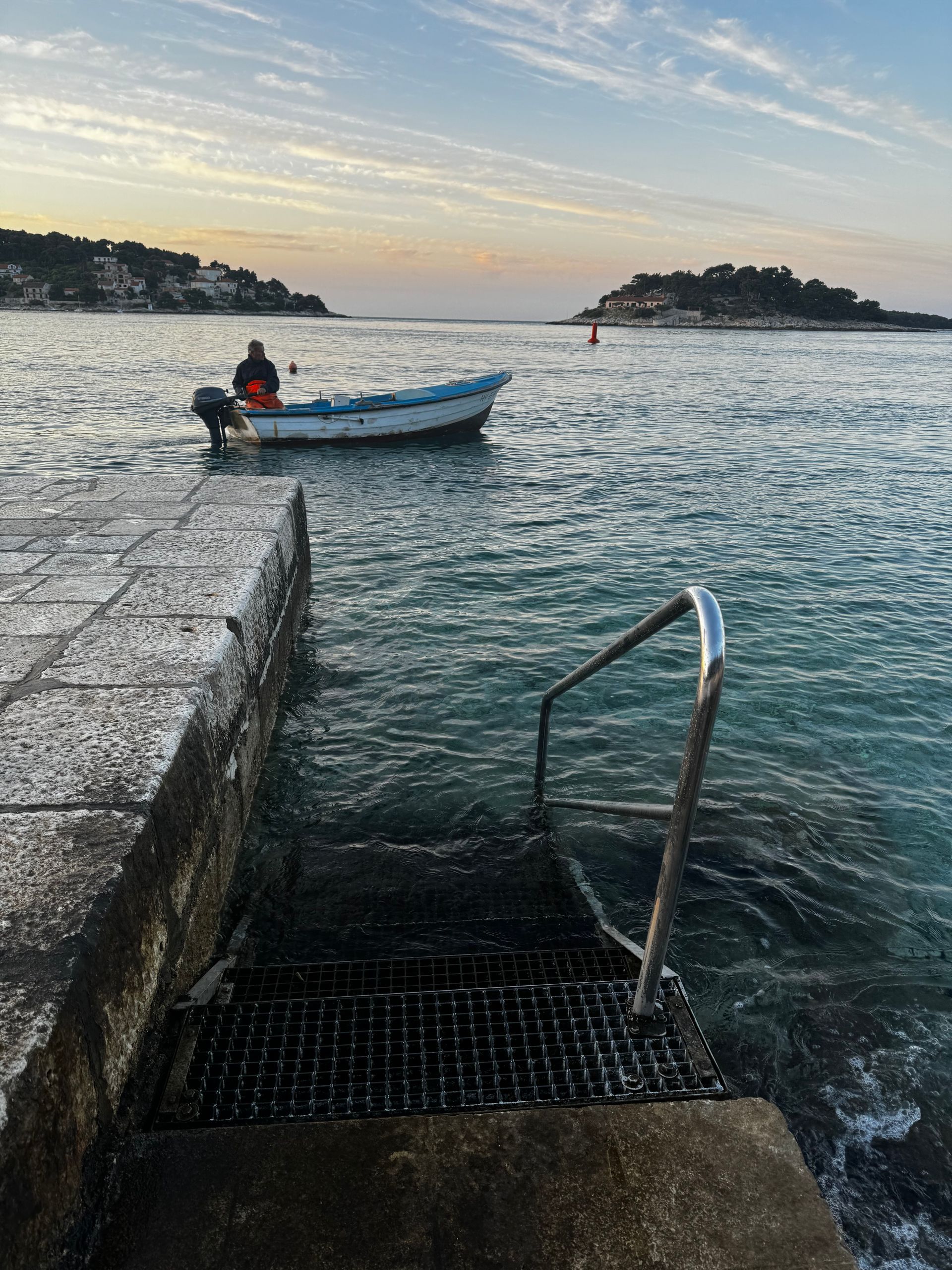 Concrete pier with metal ladder leads to water, boat with person in distance, island, sunset sky.