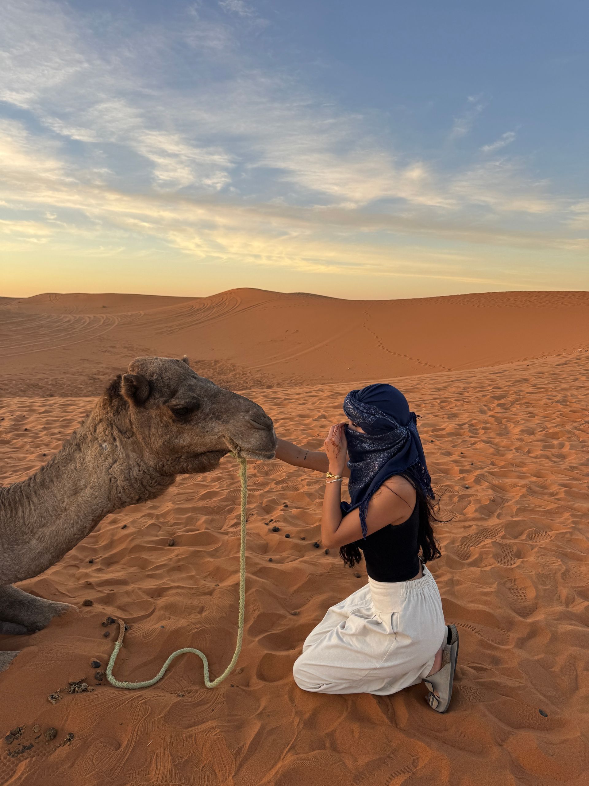 Woman kneeling, petting camel in desert at sunset. Sand dunes and orange sky in background.