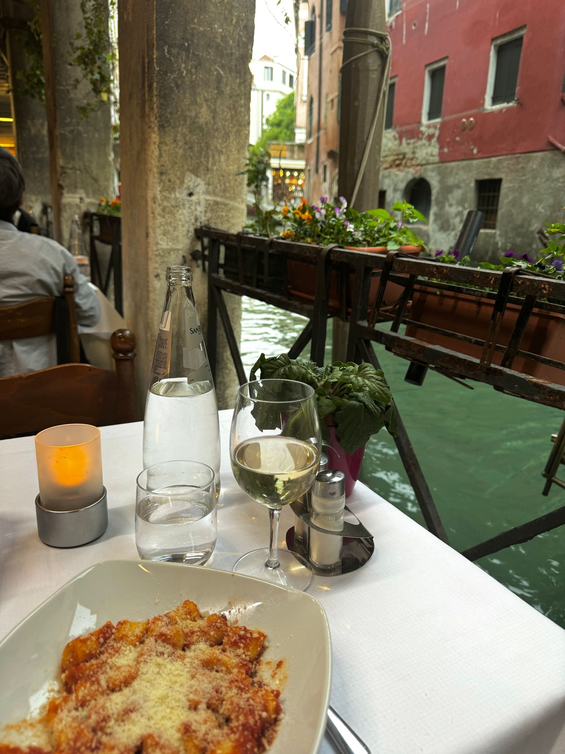 Table set for meal alongside a Venetian canal, featuring pasta, wine, water, and candle.