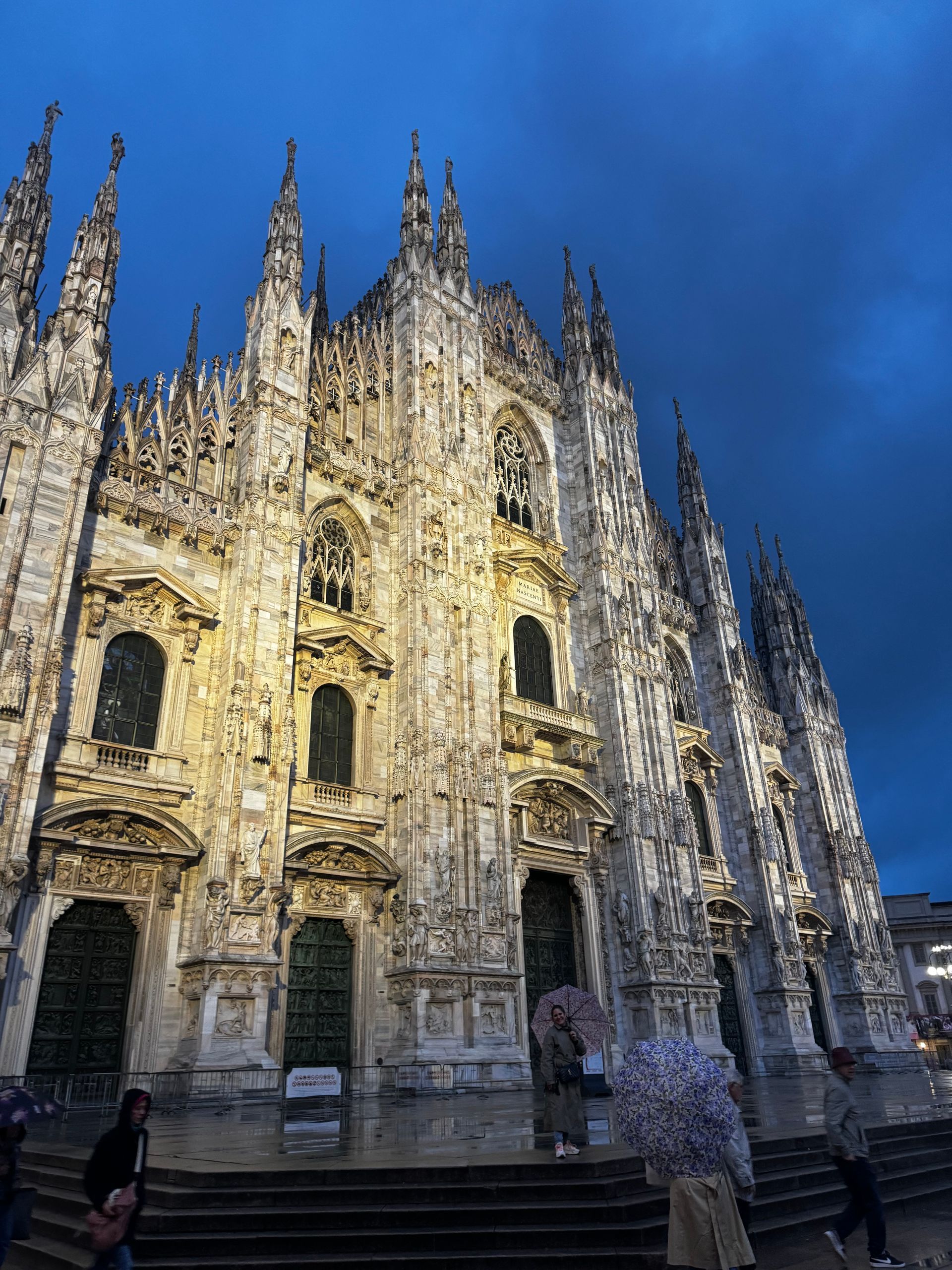 Duomo di Milano at dusk, illuminated facade against a dark blue sky, people below.