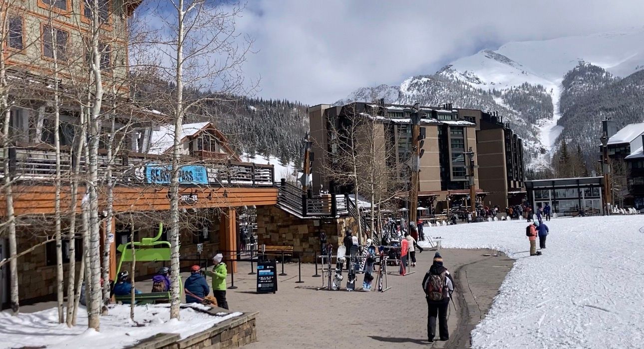 Snowy ski resort scene with buildings, skiers, and mountain backdrop.