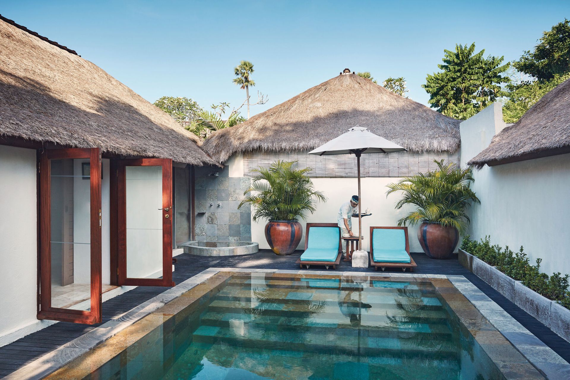 A small swimming pool is surrounded by thatched-roofed villa in Bali.