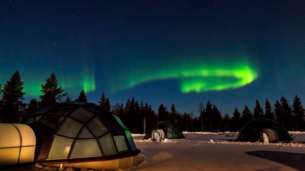 The aurora borealis is visible in the night sky over a snowy field with glass igloos in finland.