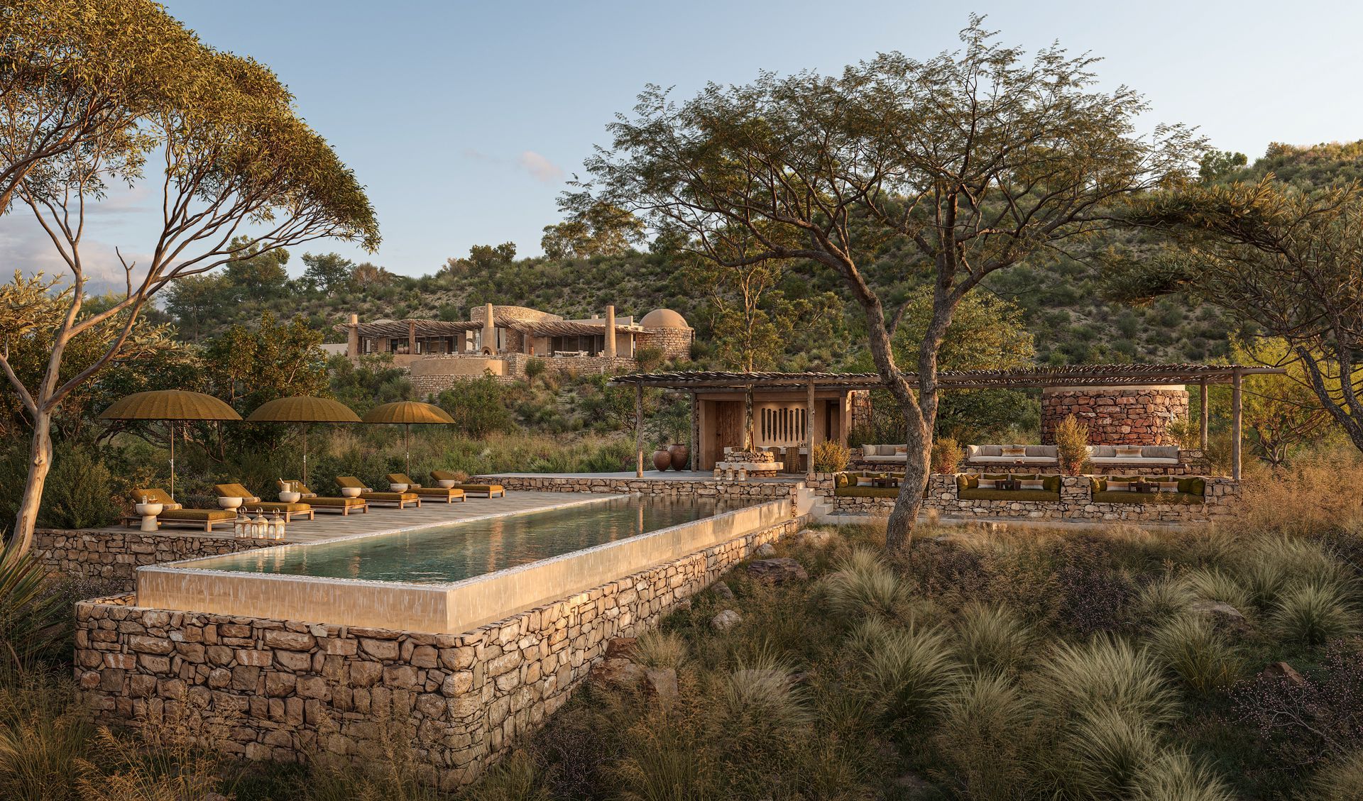 A large swimming pool is surrounded by trees and umbrellas in front of a lodge in Africa.