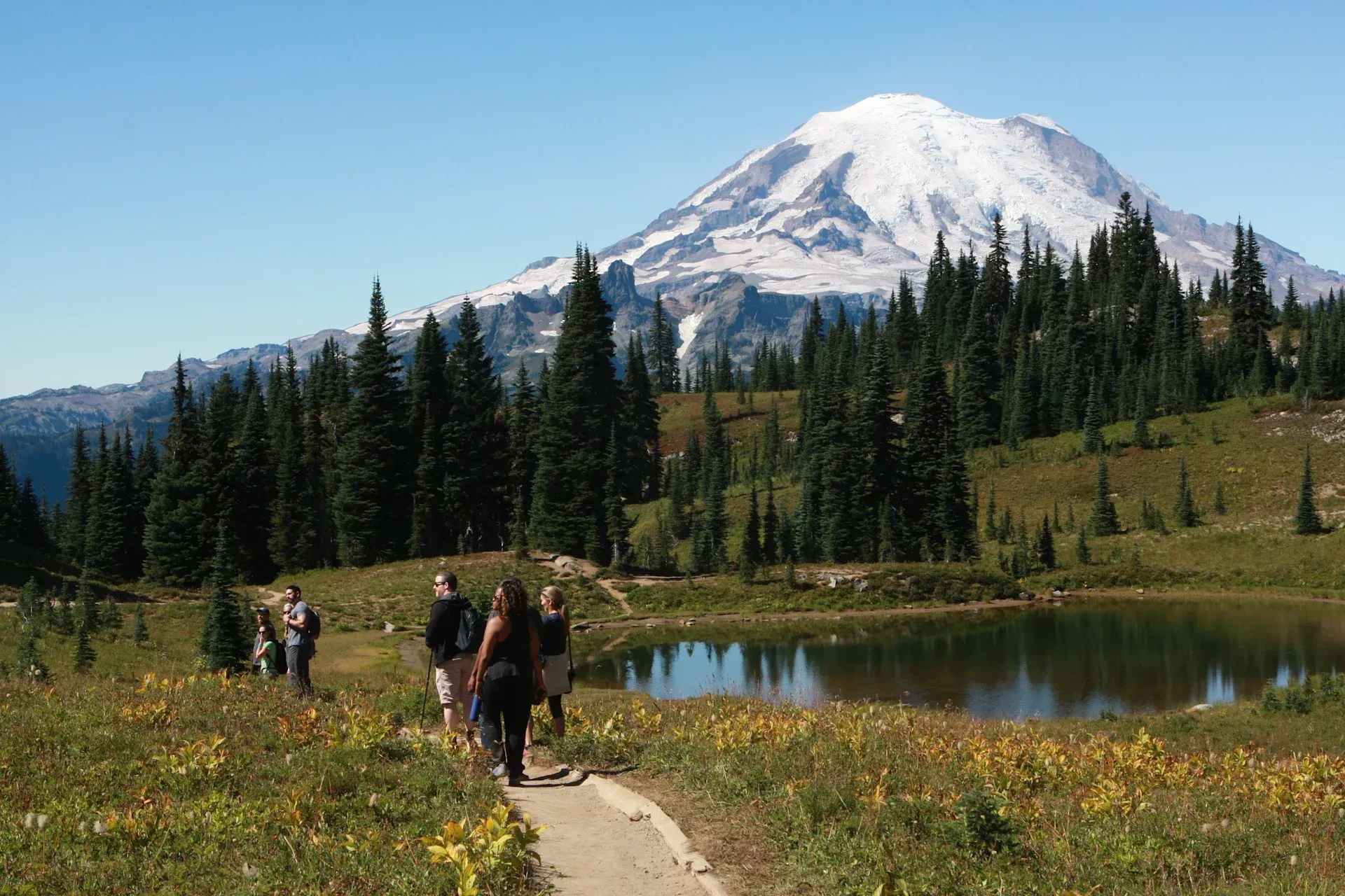 A group of people are walking along a path next to a lake with a mountain in the background.