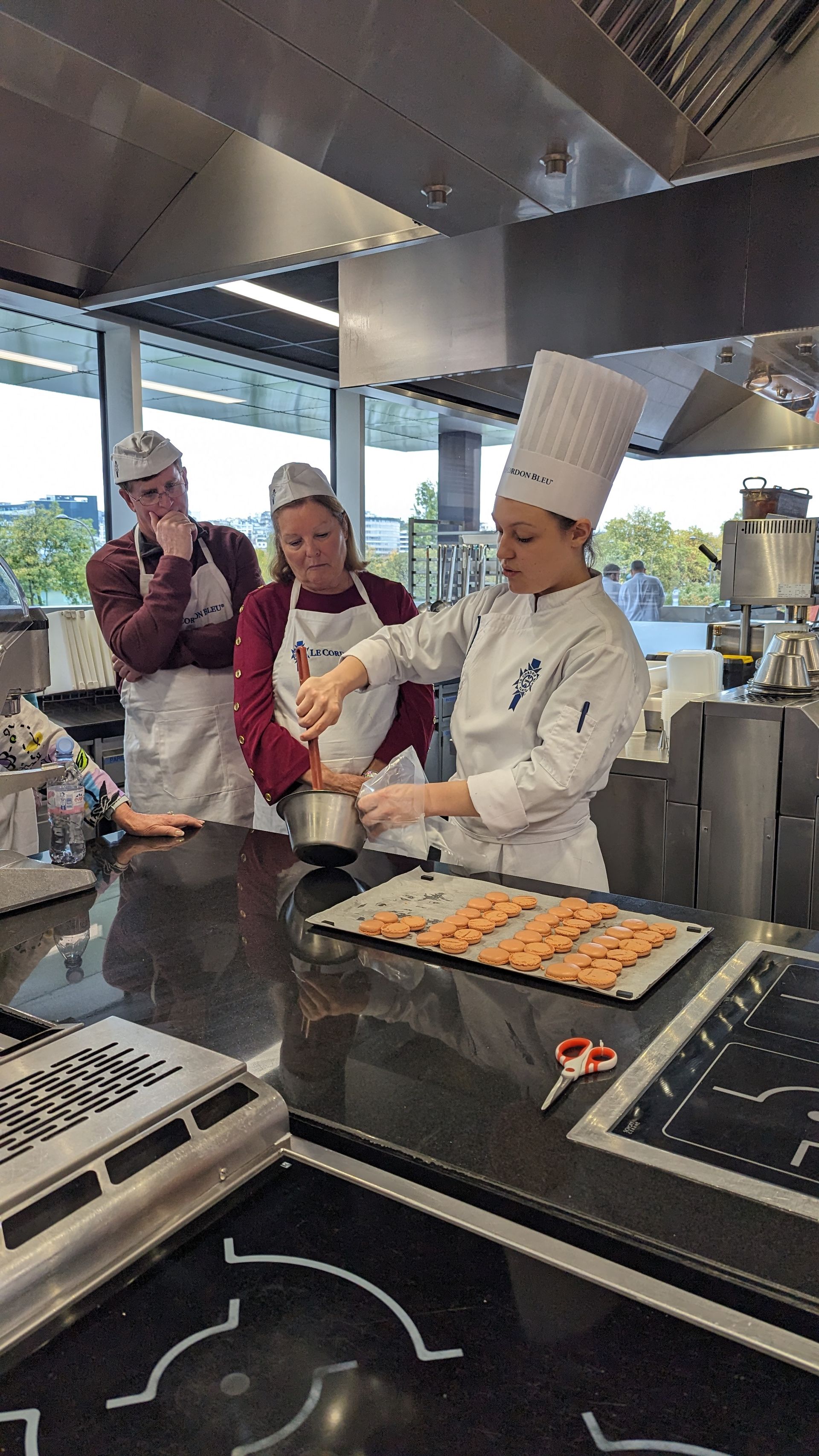 Chef demonstrating cooking technique in a kitchen, students watching. Stainless steel appliances, food preparation.