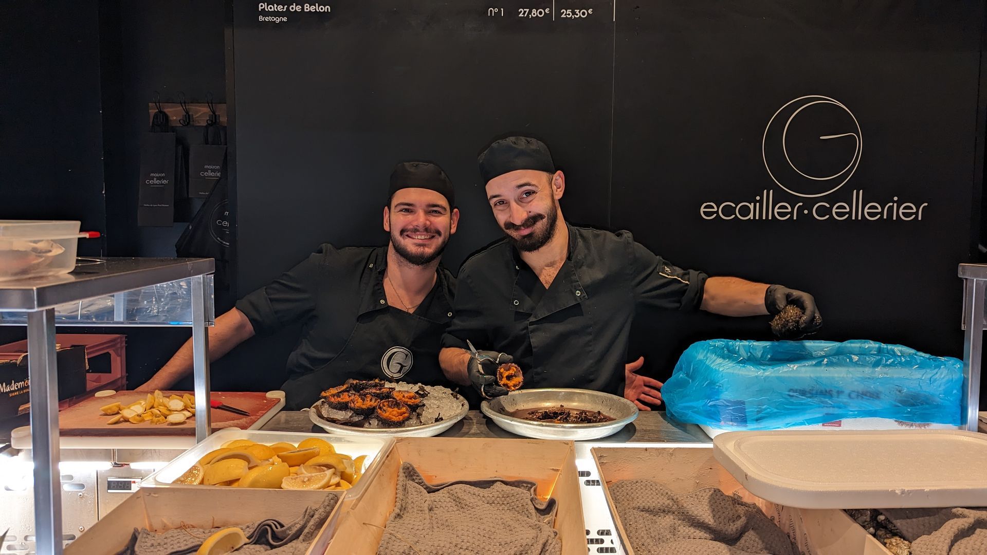 Two smiling chefs at a counter with cooked food and lemons. Black background with logo.