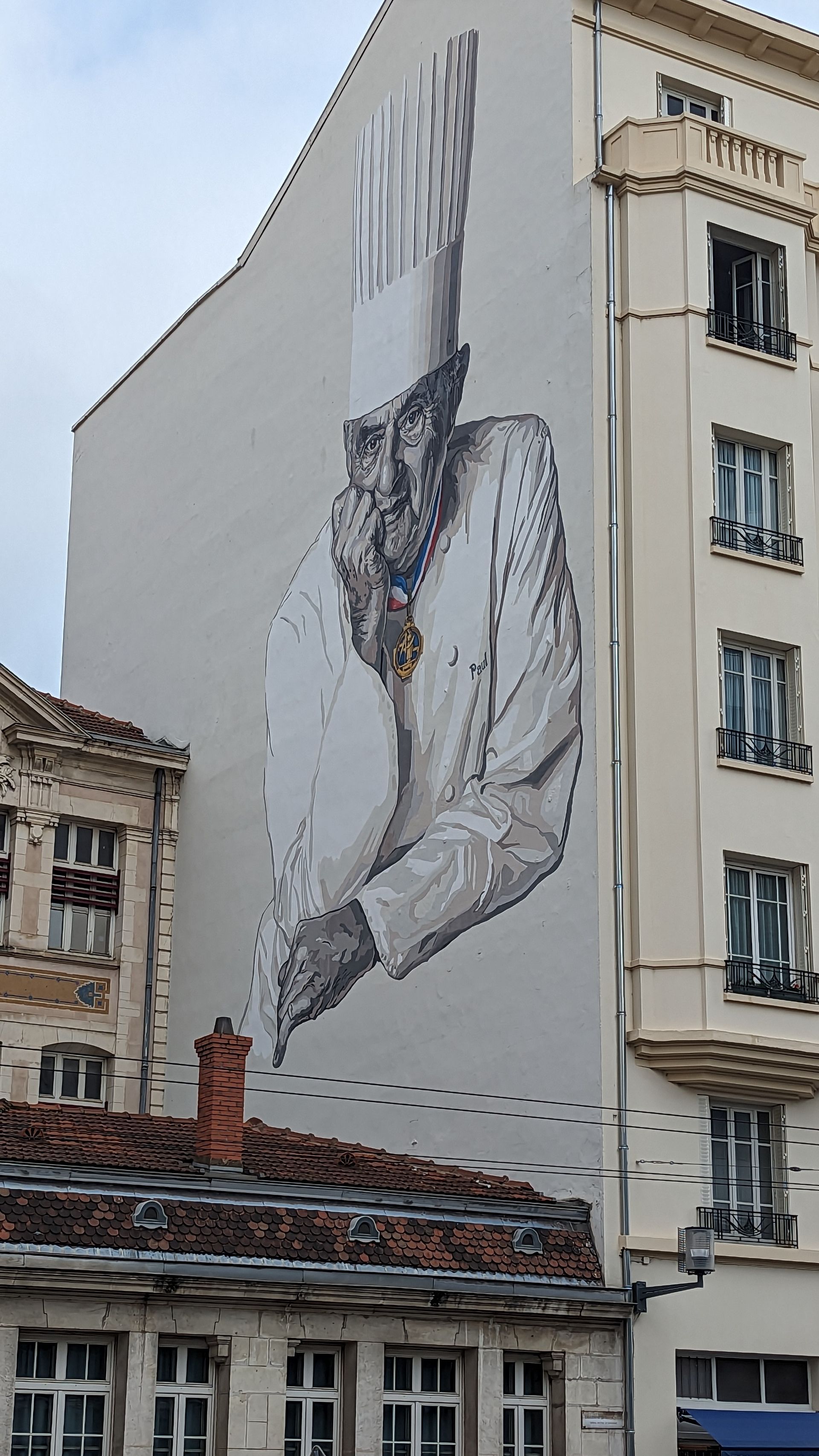 Mural on a building: Chef wearing a white jacket and hat, painted in shades of gray. Buildings surround.