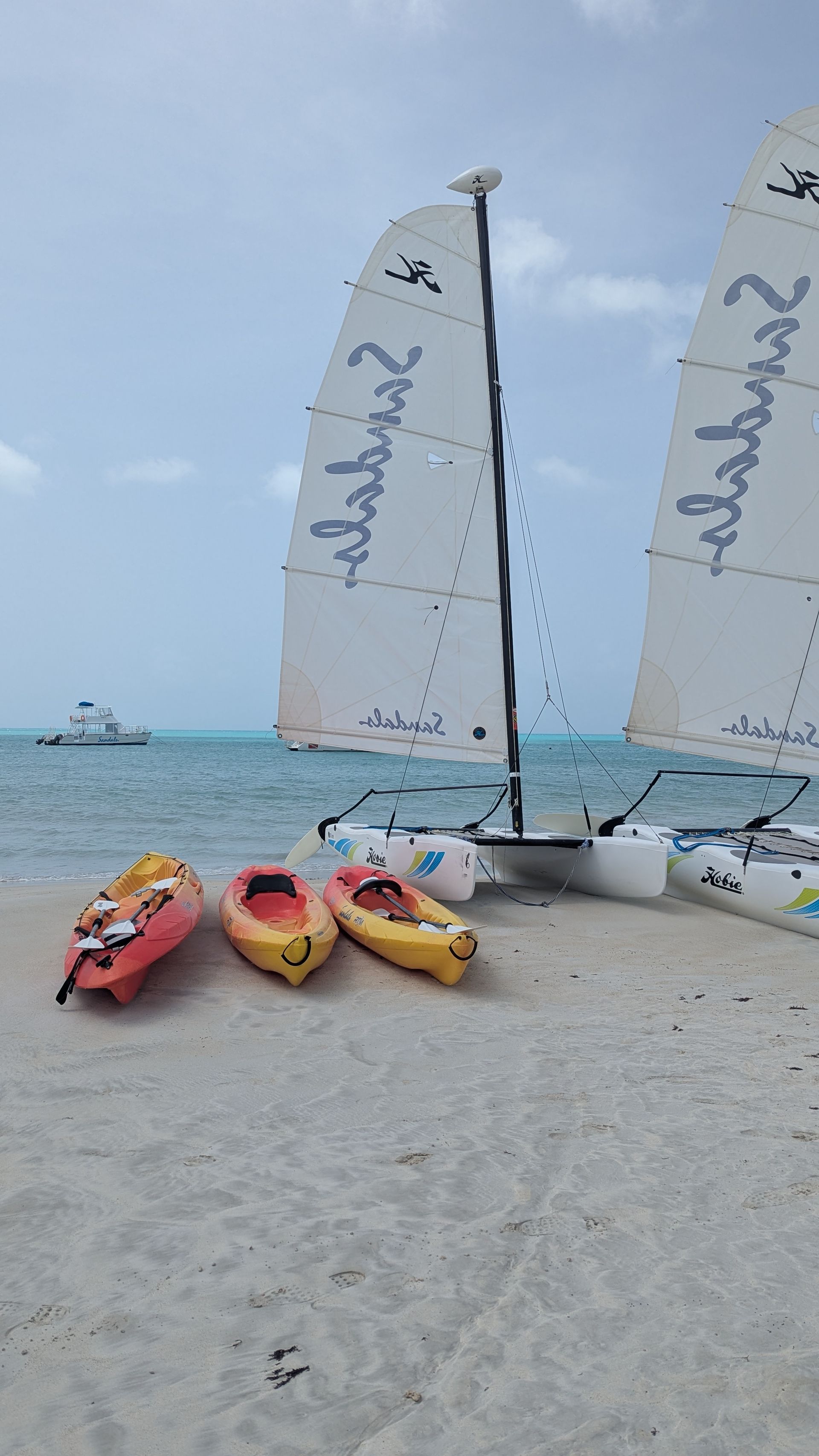 Kayaks on a sandy beach with sailboats in the background, under a cloudy sky.