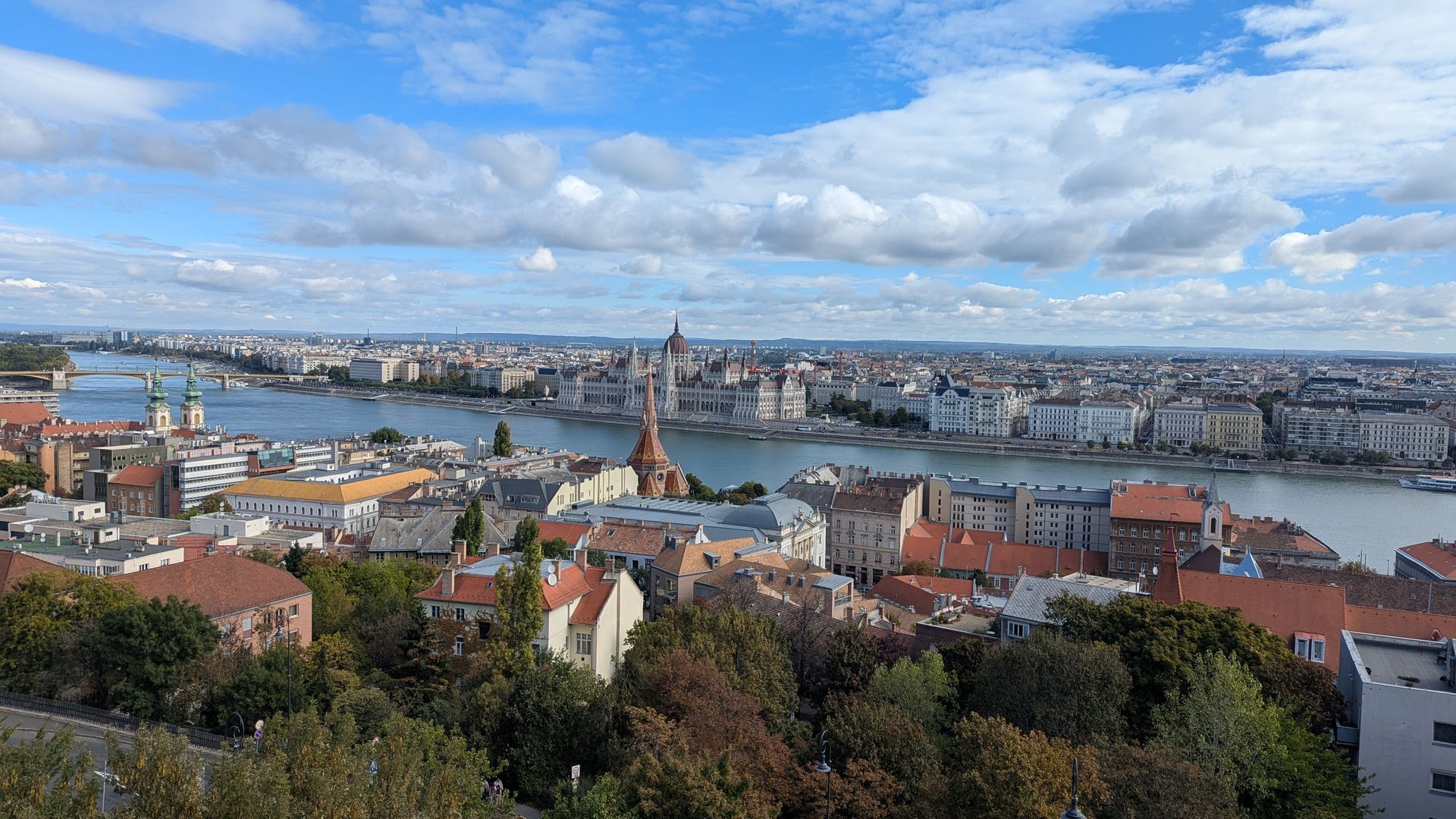 Panoramic cityscape with a river, buildings, and blue sky with clouds.