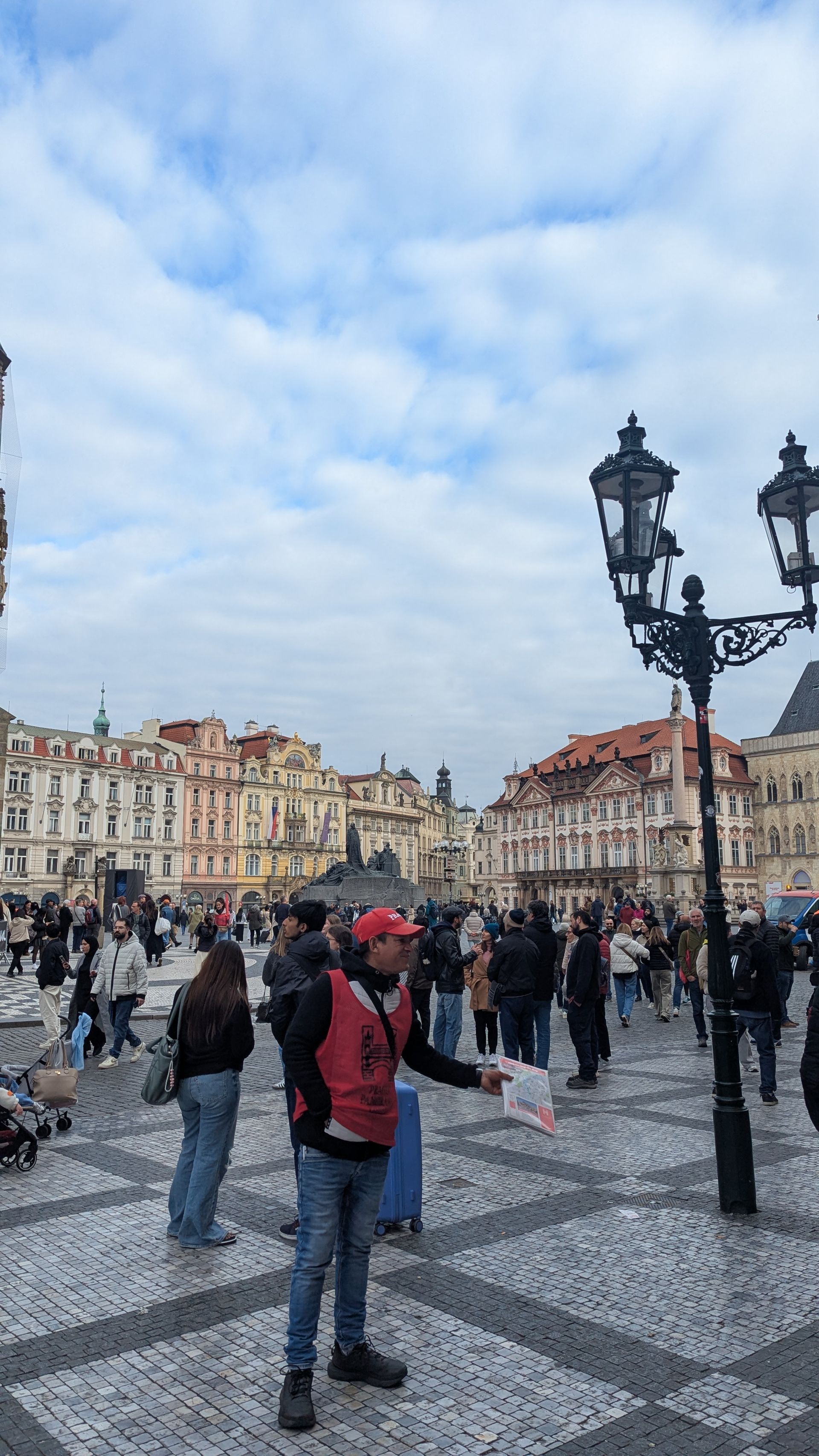 Man in red vest gestures toward crowd in European square; buildings in background.