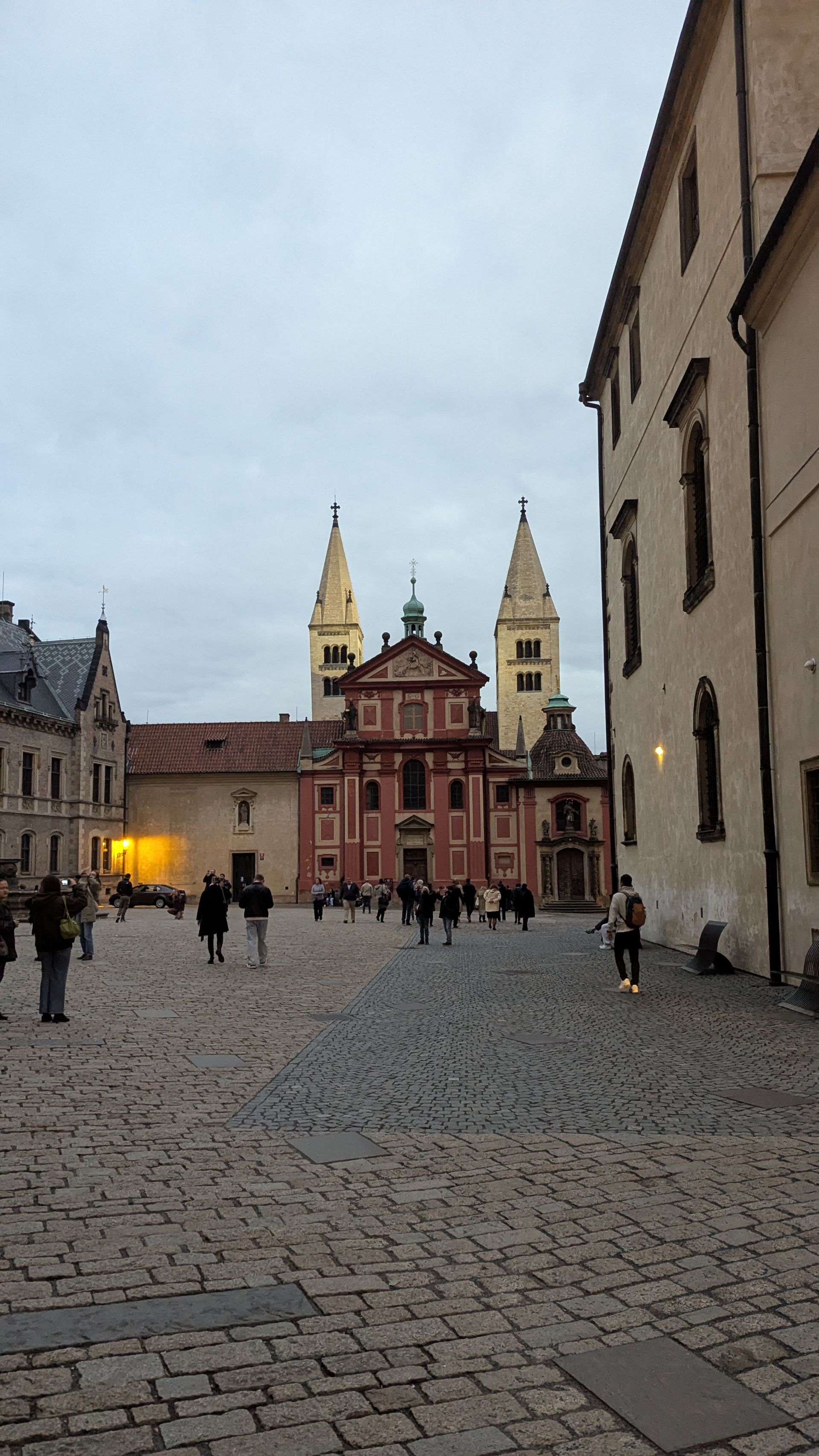 Cobblestone square with people walking toward a red church with two tall, yellow spires. Gray sky.