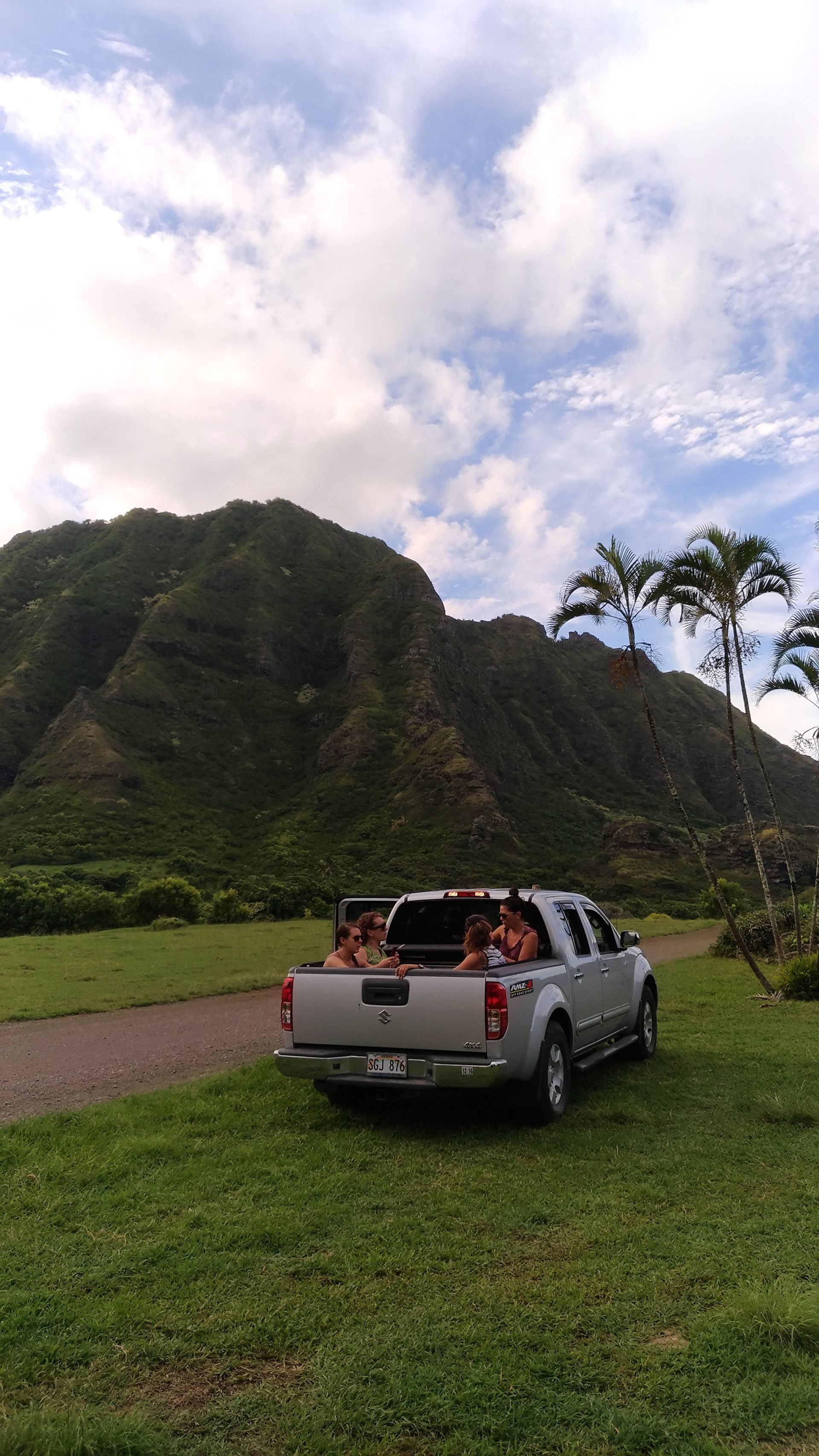 Silver pickup truck with passengers, parked on grass near mountain. Cloudy sky.