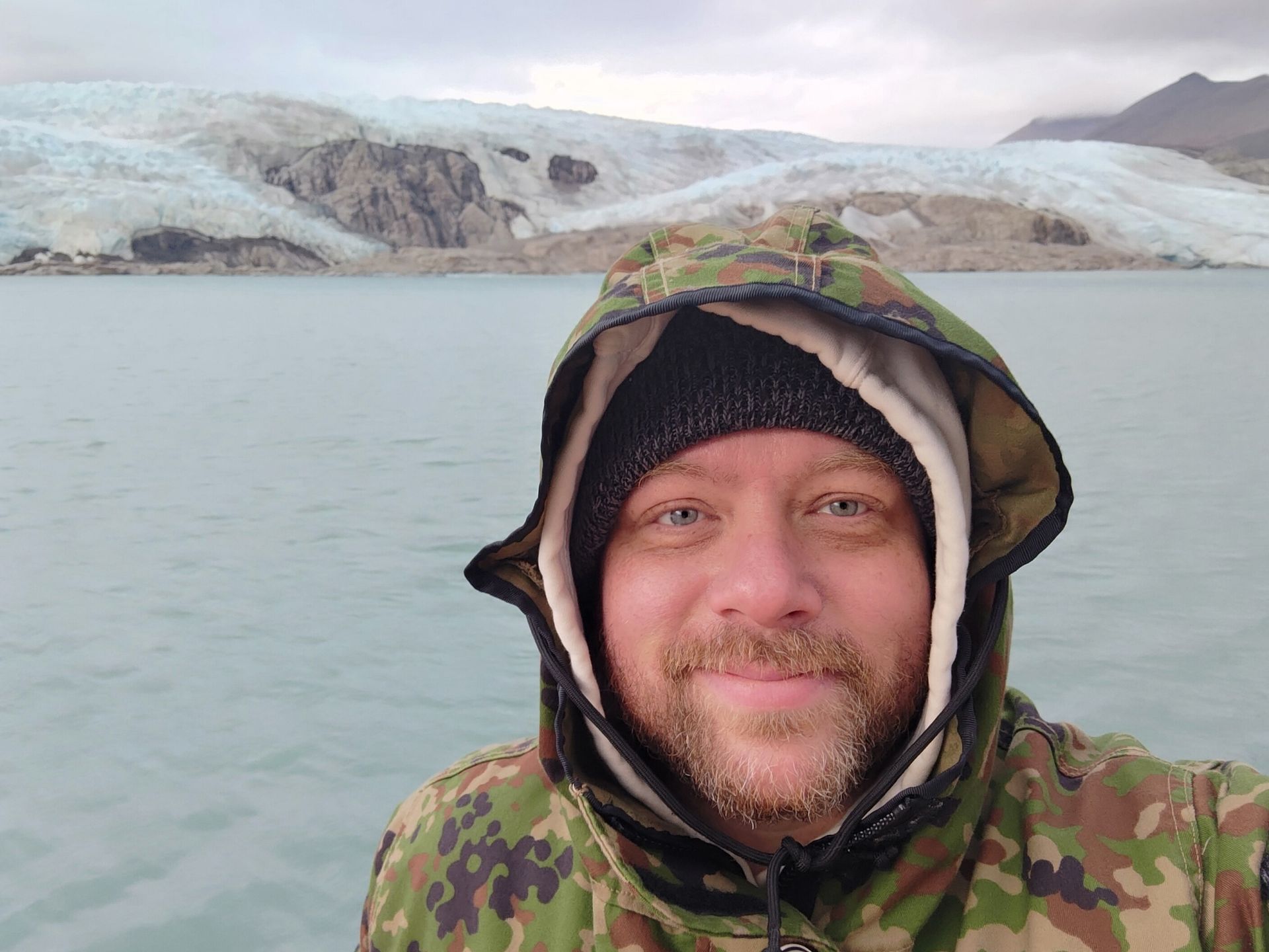 A man in a camouflage jacket is standing in front of a lake.