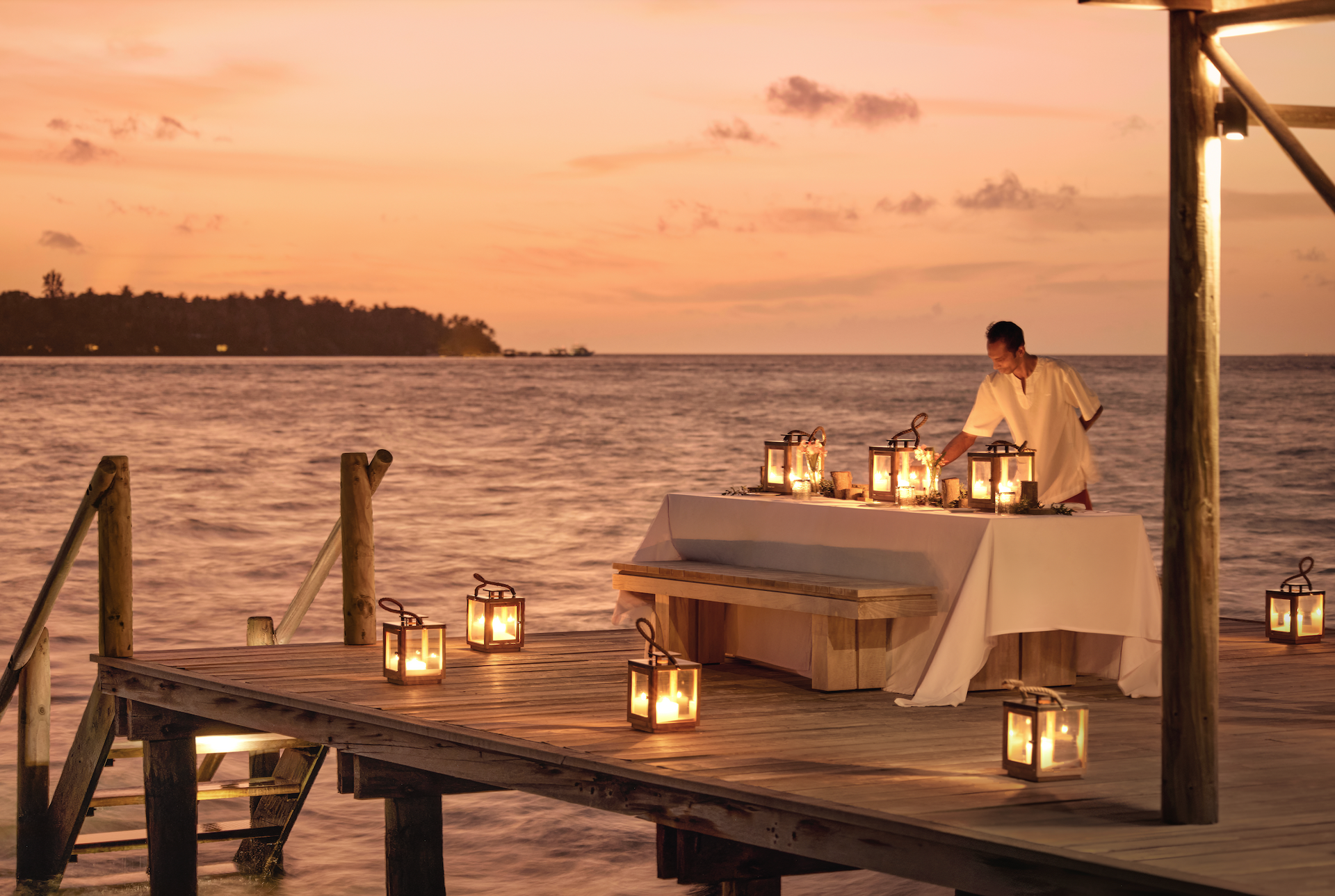 A man is preparing a table on a dock overlooking the ocean at sunset in the Maldives at como resort. 