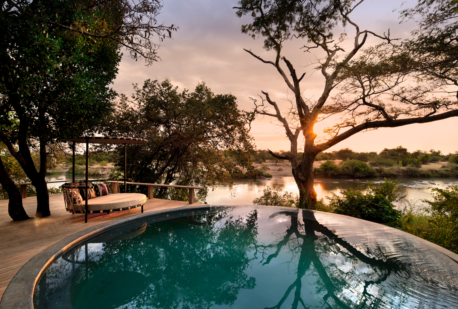 A large swimming pool surrounded by trees and a river at a lodge in Africa.