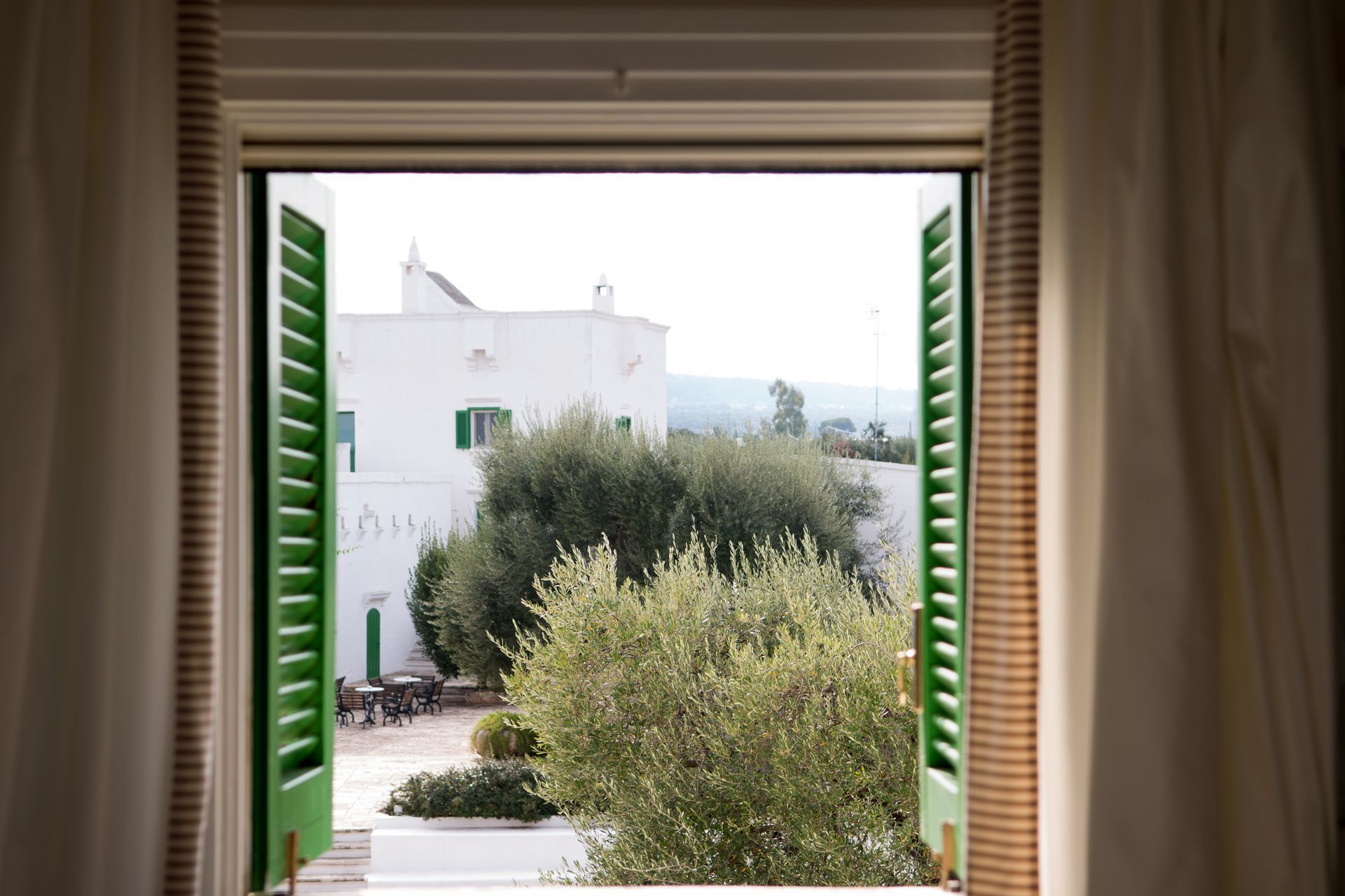 An open window with green shutters looking out to a white house at Il Melograno Hotel in Puglia, Italy.