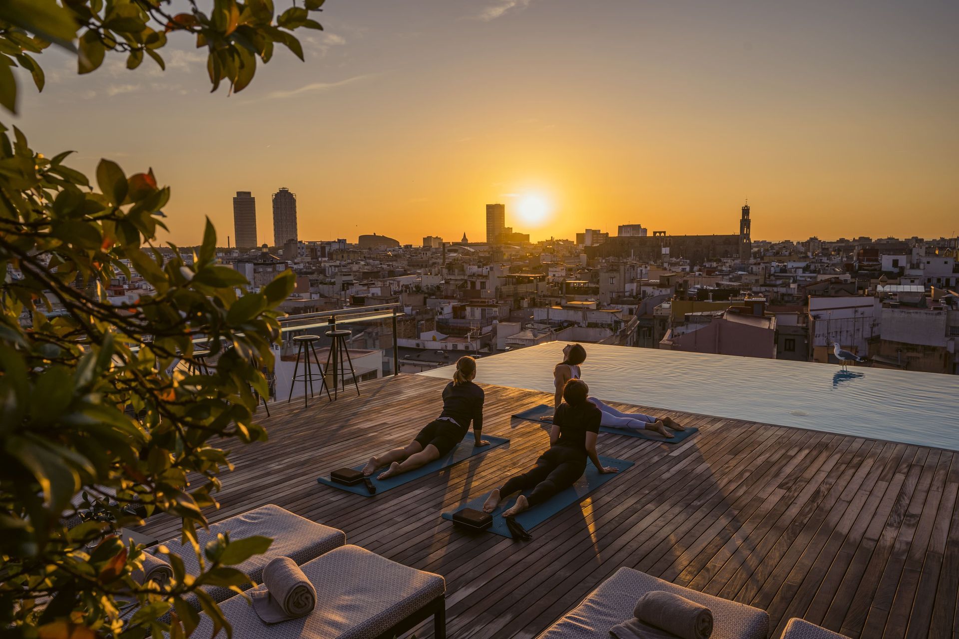 A group of people are doing yoga on a rooftop swimming pool at sunset at Grand Hotel Central in Barcelona, Spain.