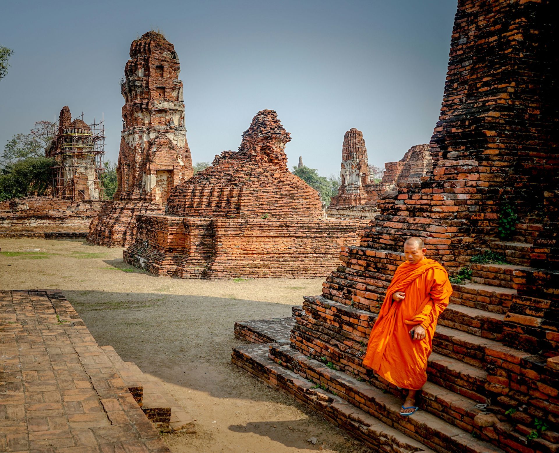 A monk in an orange robe is standing on a set of stairs in front of the ruins of Ayutthaya, Thailand.