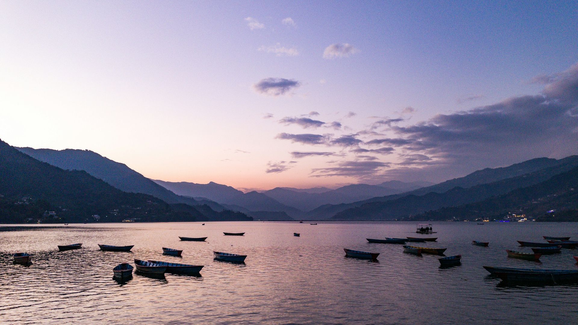 Boats on calm lake at dusk; mountains in background in Pokhara, Nepal.