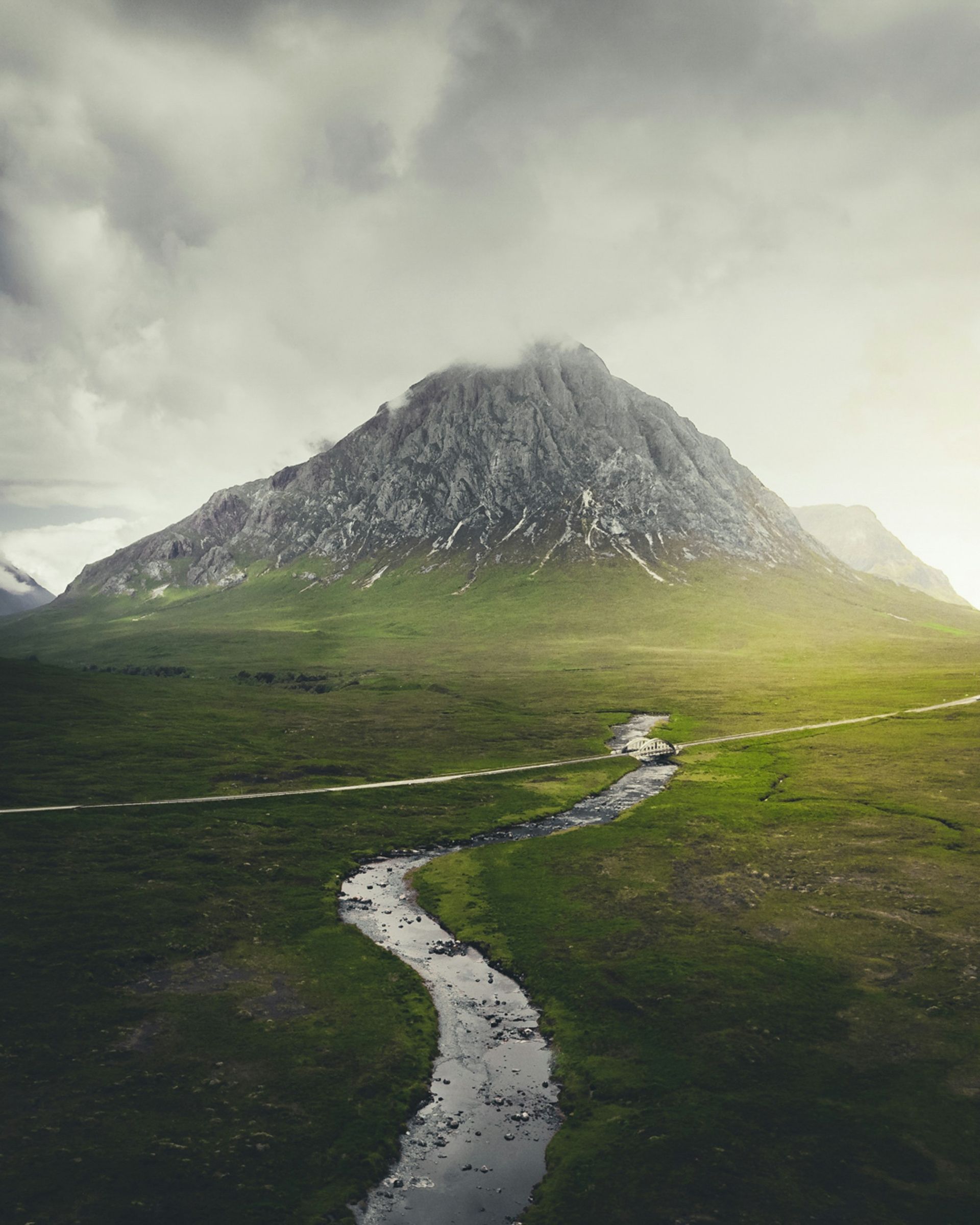 Mountain rises over a green valley, with a winding stream. Cloudy sky in Glencoe, Scotland.