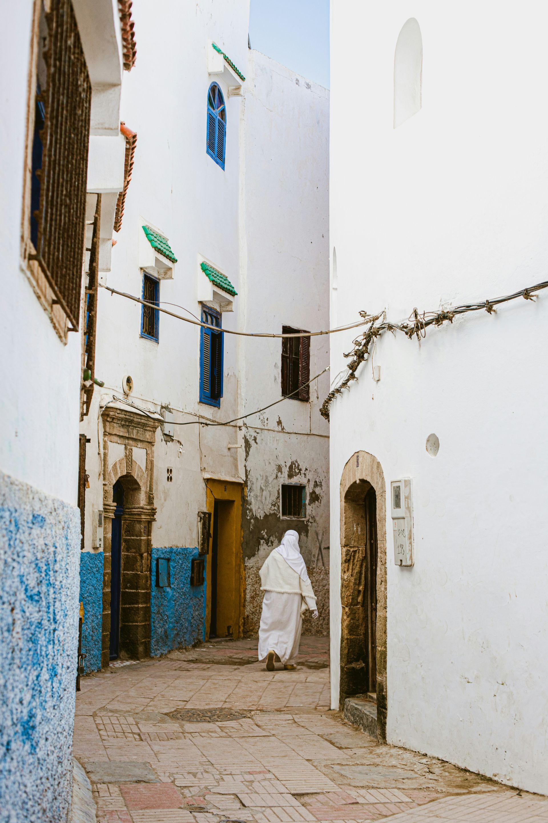 A man in a white robe is walking down a narrow street between two white buildings in Essaouira, Morocco.