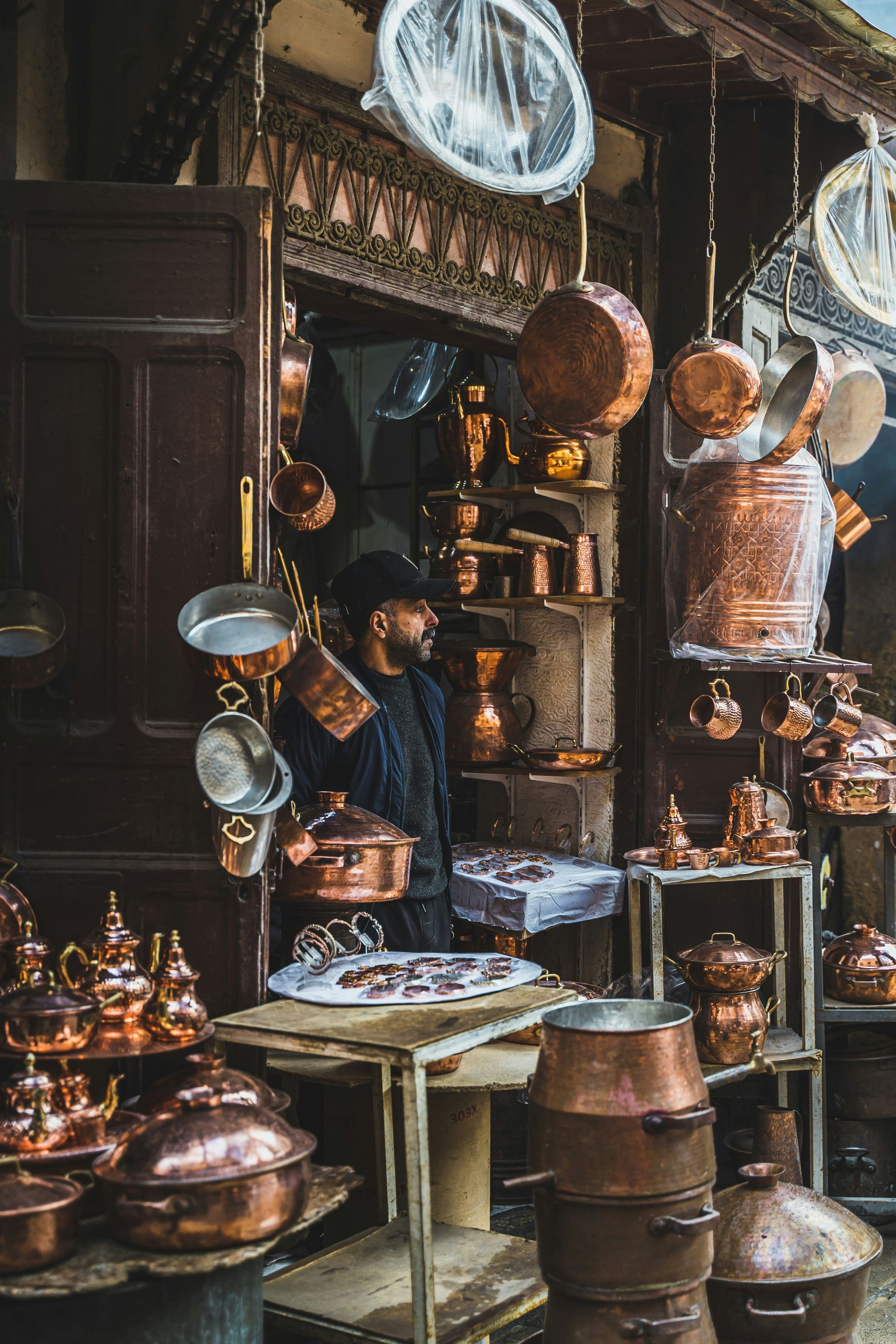 A man is standing in front of a store filled with copper pots and pans in Fes, Morocco.