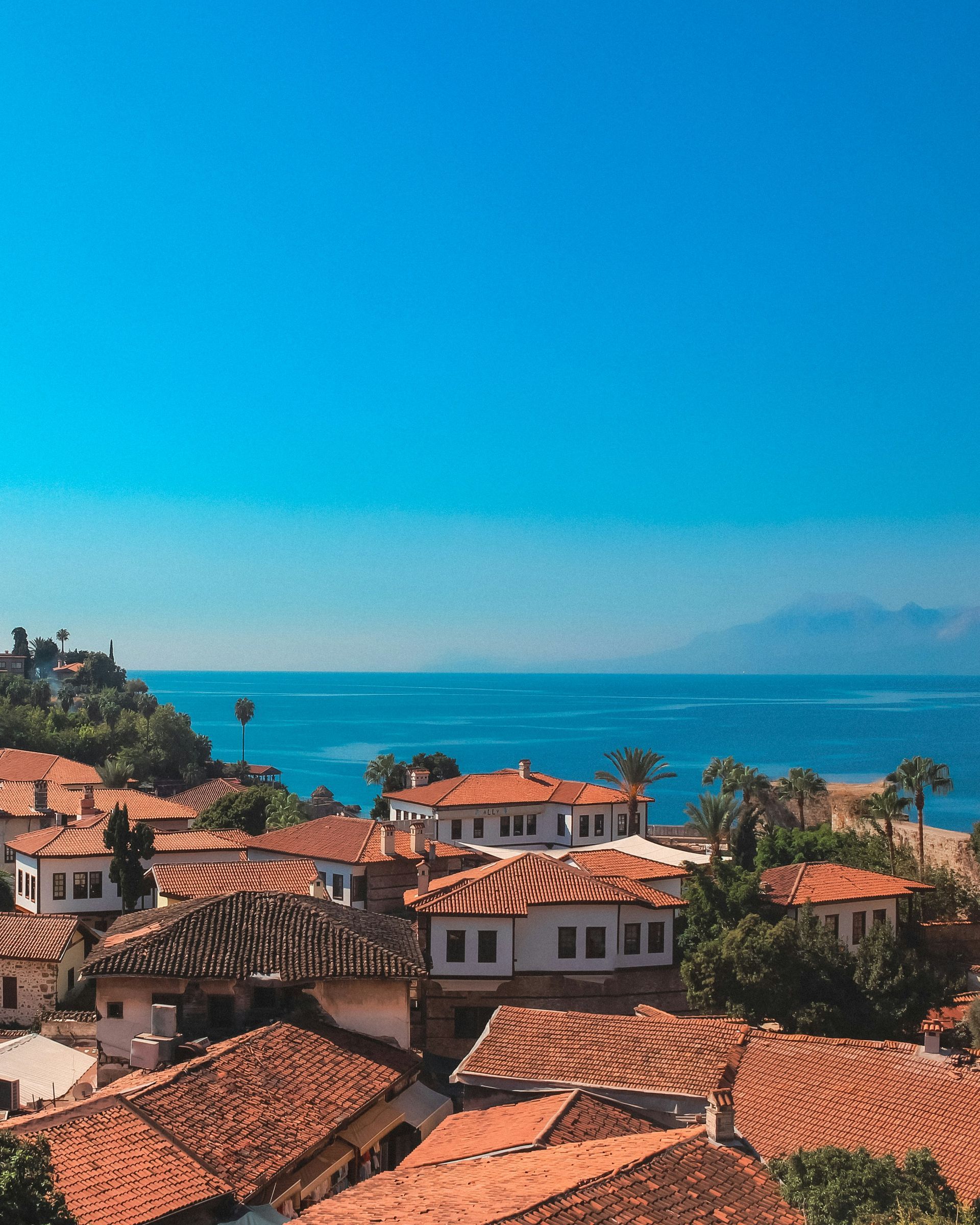 Terracotta rooftops overlook a turquoise sea under a bright blue sky in Antalya, Turkey.