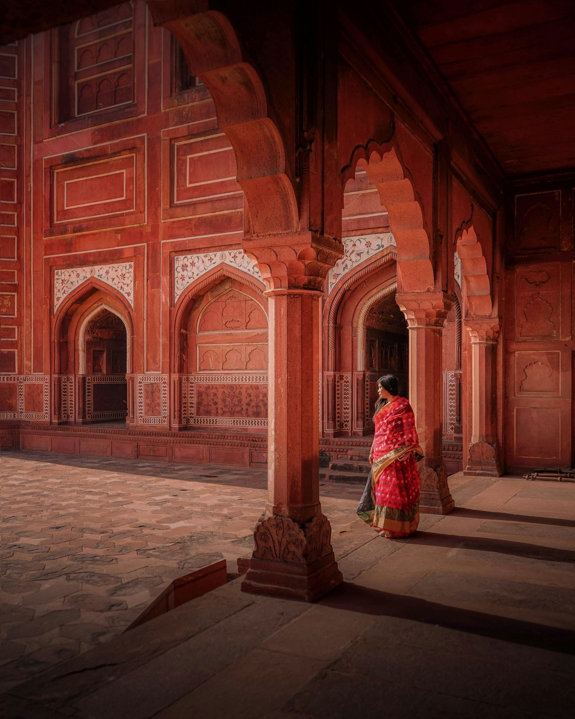 Woman in red sari walks through a red-toned archway in Agra, India.