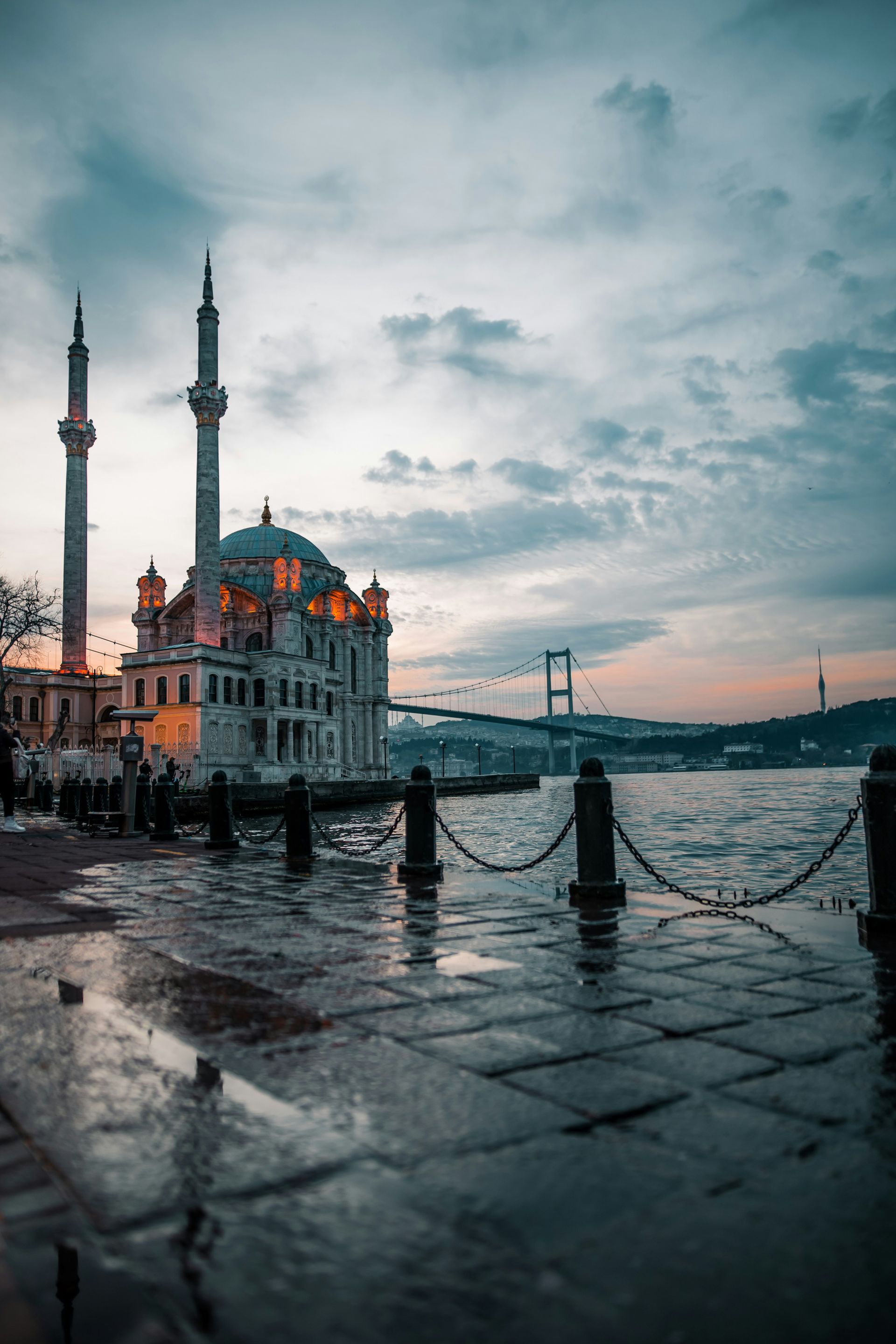 Mosque on a waterfront with two minarets, wet cobblestone, and a bridge in Istanbul, Turkey.