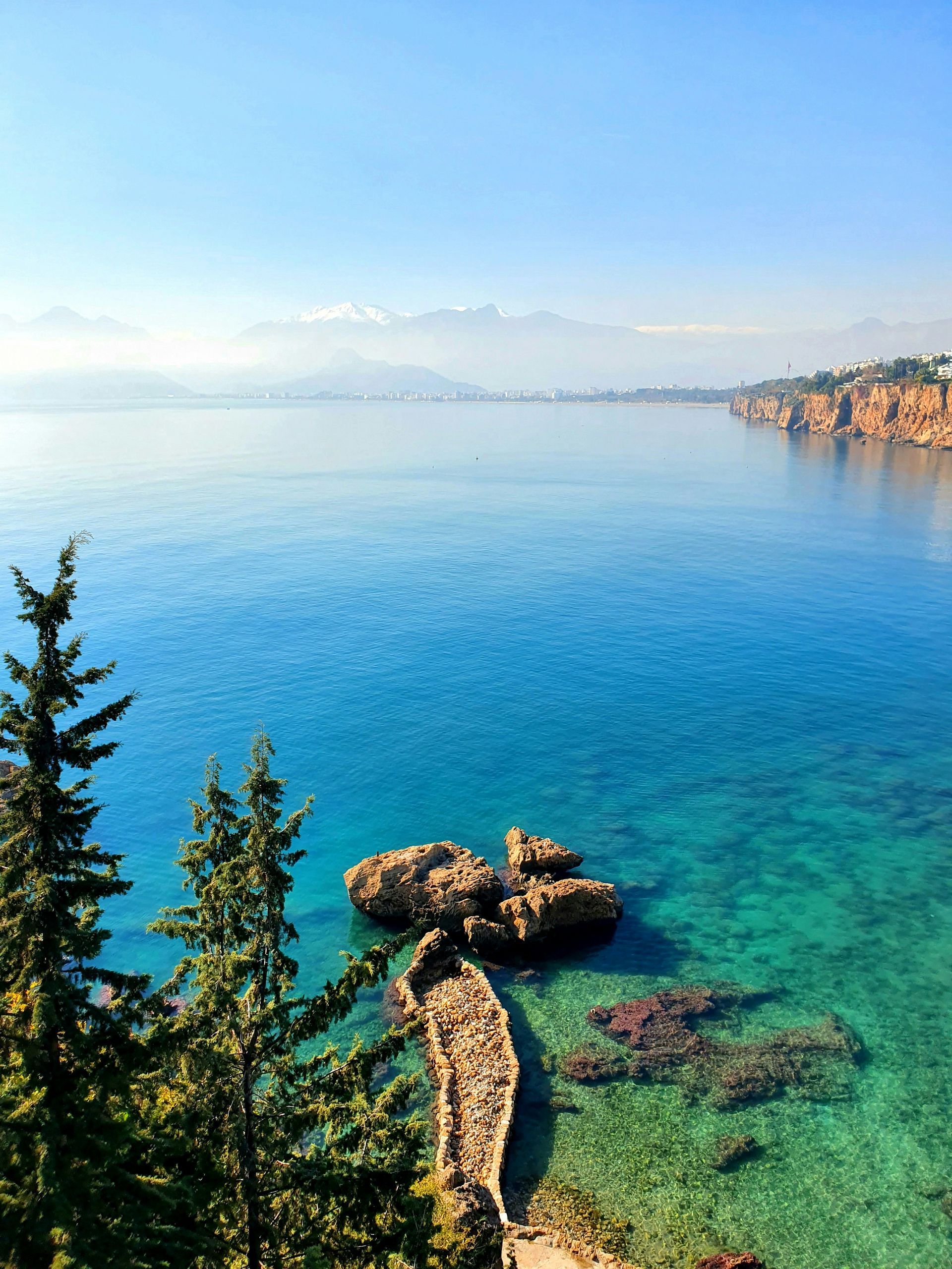 Turquoise sea, rocky shore, stone path, cliffs, mountains in distance in Antalya, Turkey.