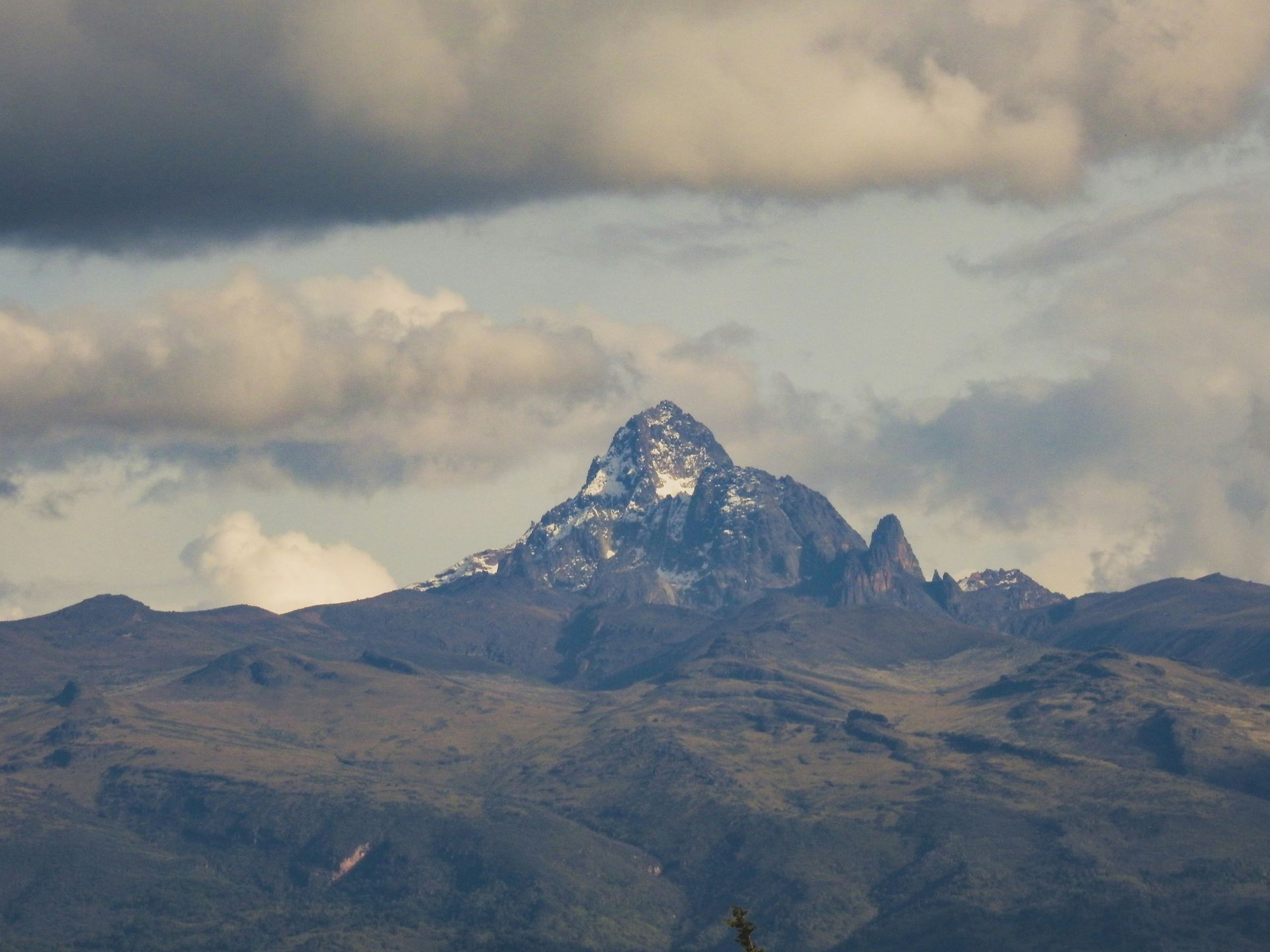 Snow-capped Mount Kenya rises from rugged terrain beneath cloudy sky.