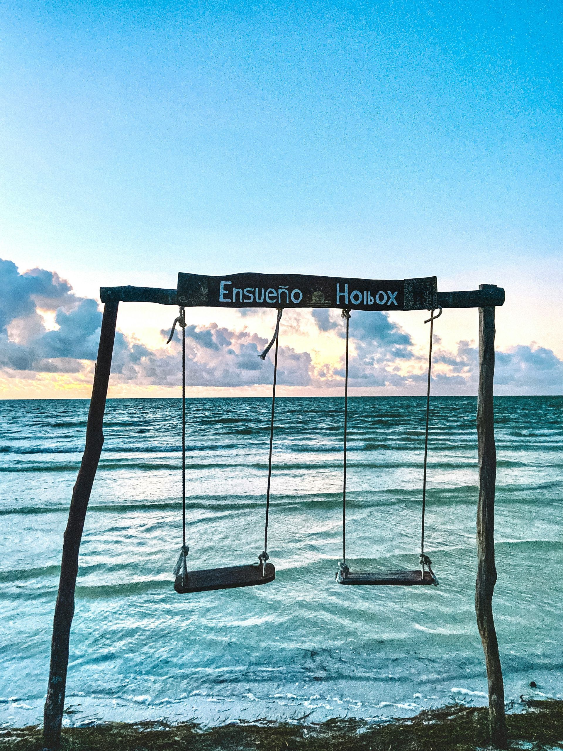 Two swings on a beach at sunset, with a sign reading 
