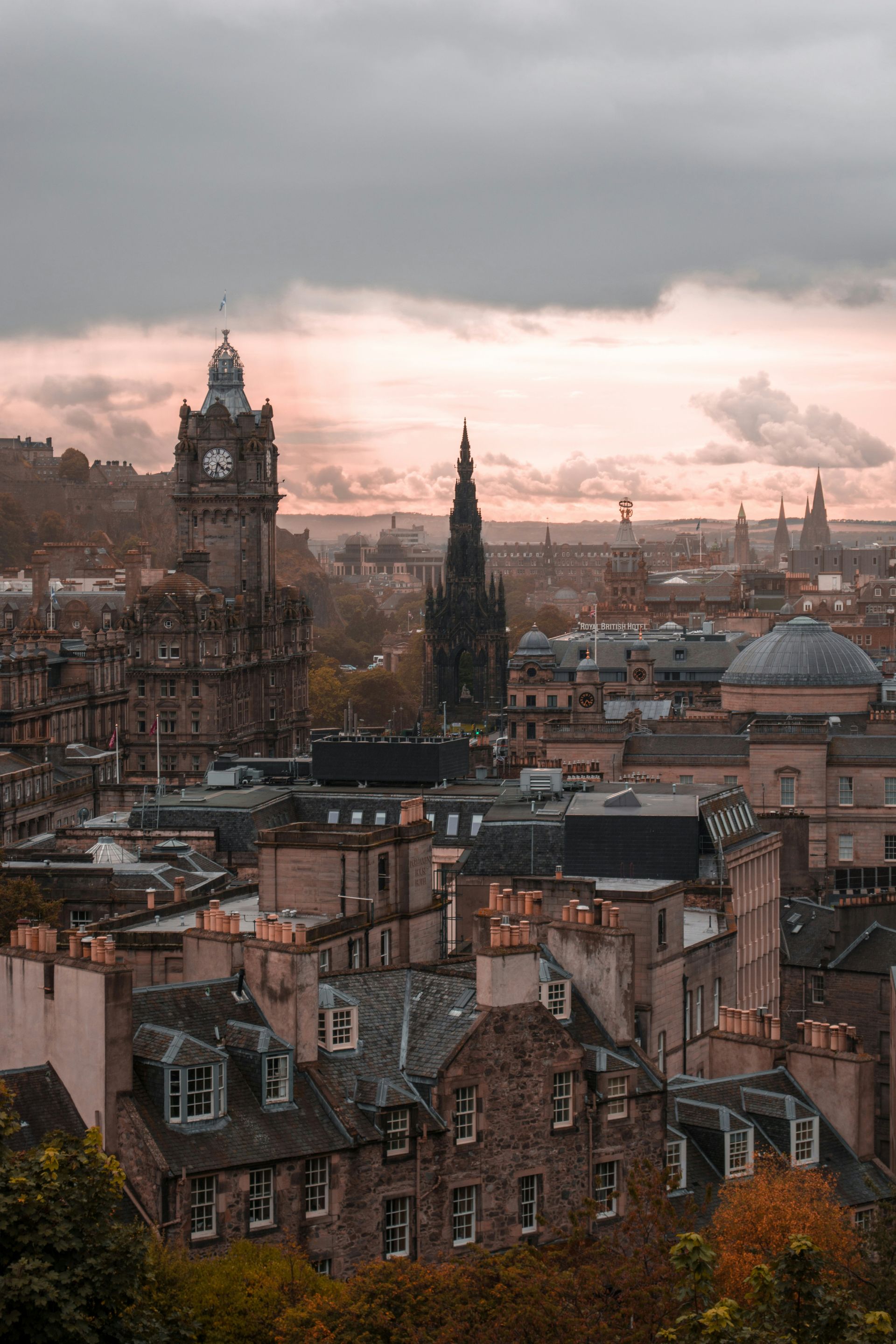 Cityscape of Edinburgh with clock tower and spires under an overcast, pink-tinged sky.